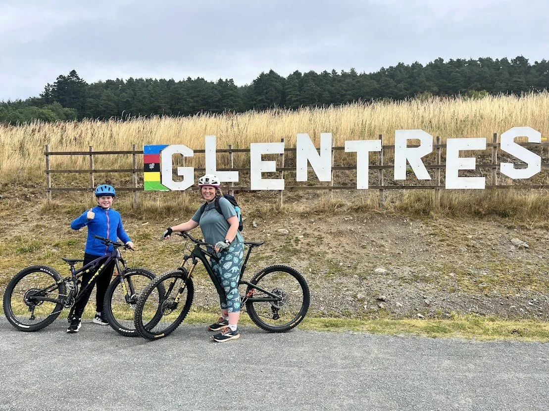 Two people with bicycles standing in front of a sign that says 'GLENTRESS' with colorful flag-like decorations, on a grassy hill under a cloudy sky.