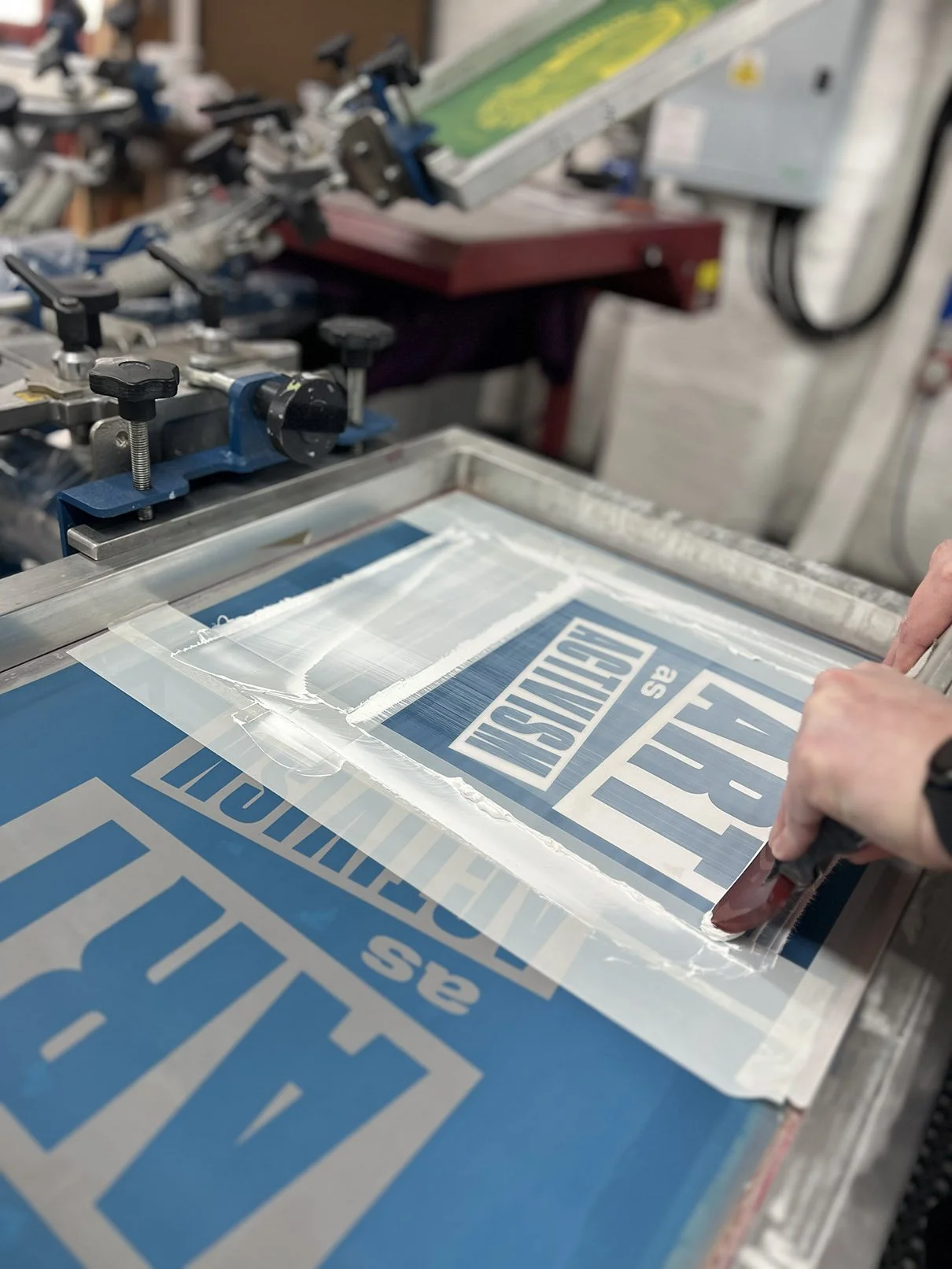A person's hands pressing a printing squeegee down on a screen printing frame, creating a custom press sign with blue and white ink.