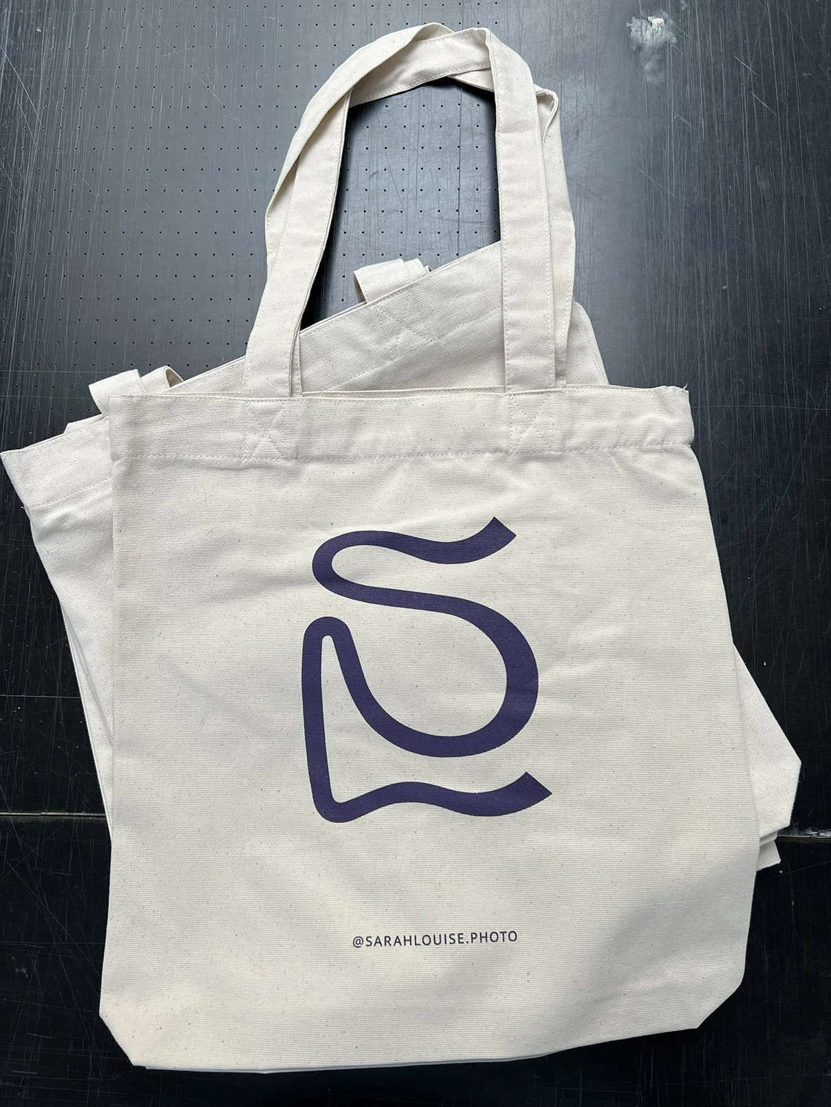 White tote bag with dark purple abstract logo and Instagram handle '@SARAHLOUISE.PHOTO' printed on it, resting on a dark surface.