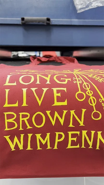 Close-up of a red bag with yellow text reading "Long Live Brown Wimpenny Brown" and a yellow decorative design, placed near a black and gray object.