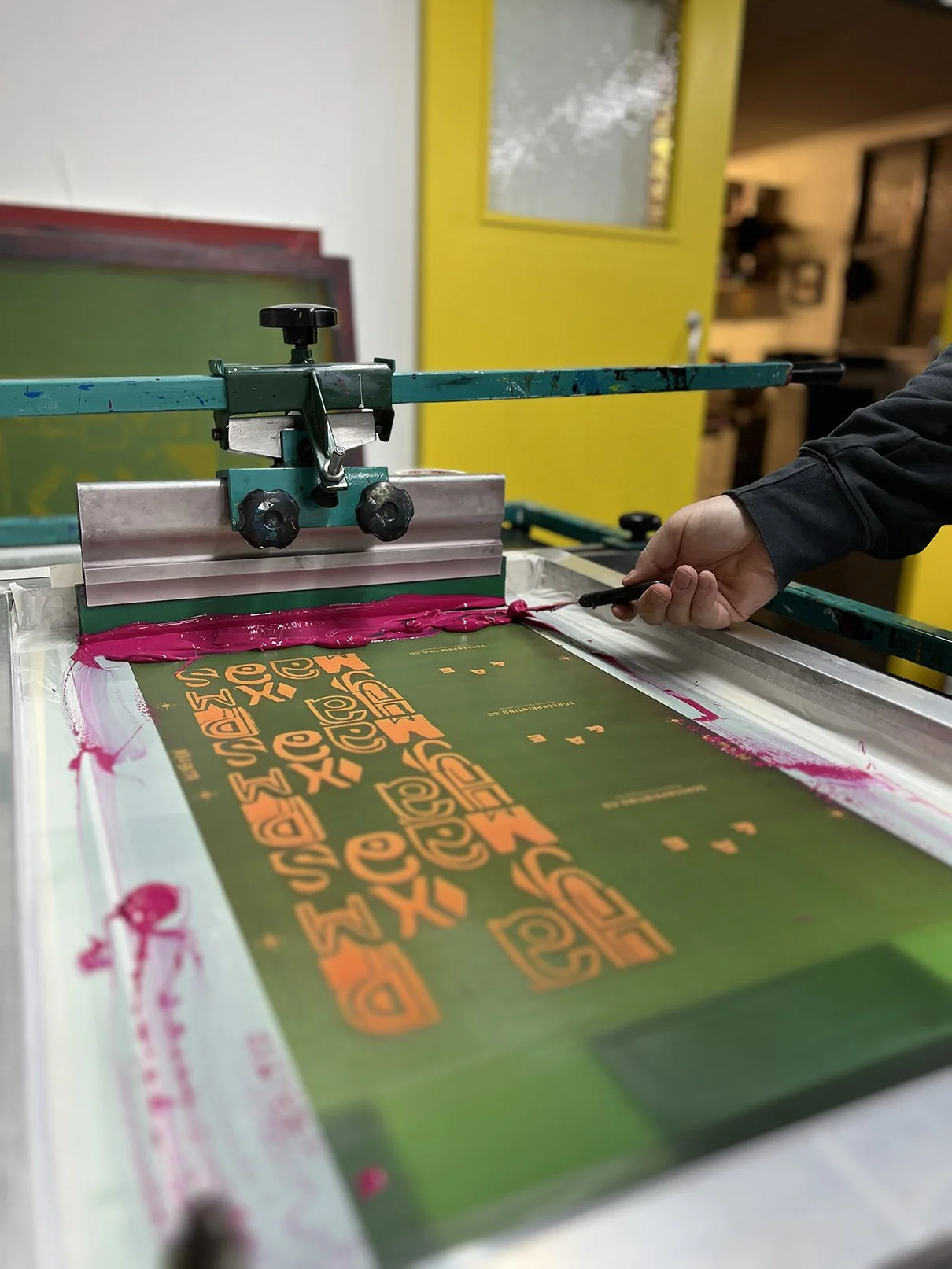 Hand screen printing cards on a flatbed printing press with orange type  onto a green surface using a screen printing frame and pink ink.