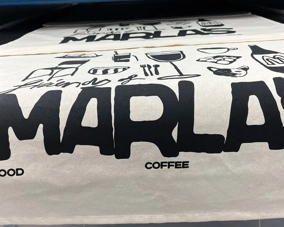 A natural cotton tea towel with black text and illustrations related to food, coffee, and a martini drink, laid out flat.