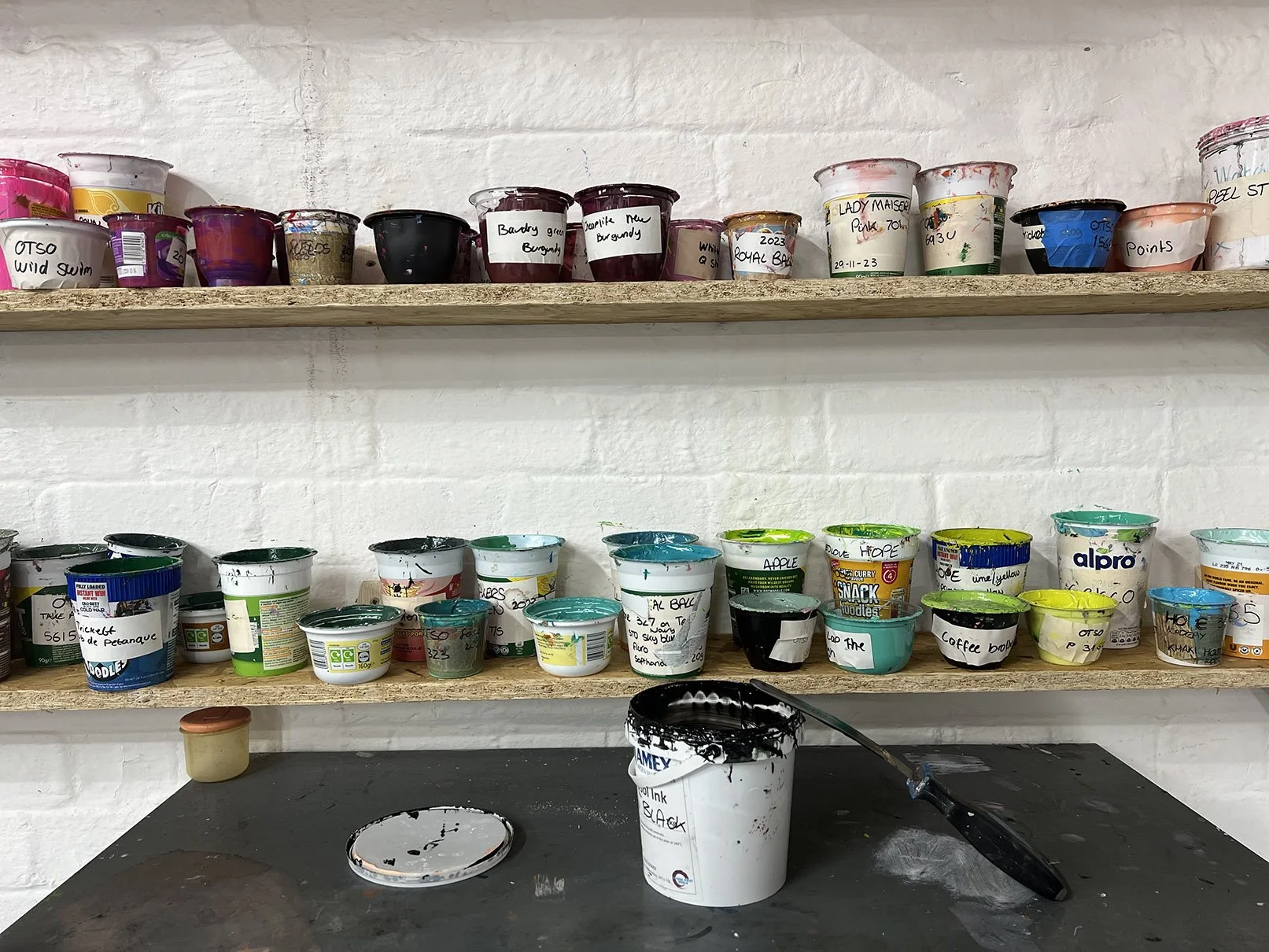 Multiple paint containers labeled with various names and colors on wooden shelves, with a black paintbrush resting in a white paint bucket on a table below.