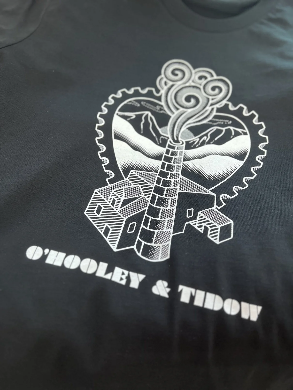 Hand screen printed graphic design featuring a factory chimney emitting smoke, with mountains in the background, surrounded by a gear, and the text ‘O’HOOLEY & TIDOW’ below.