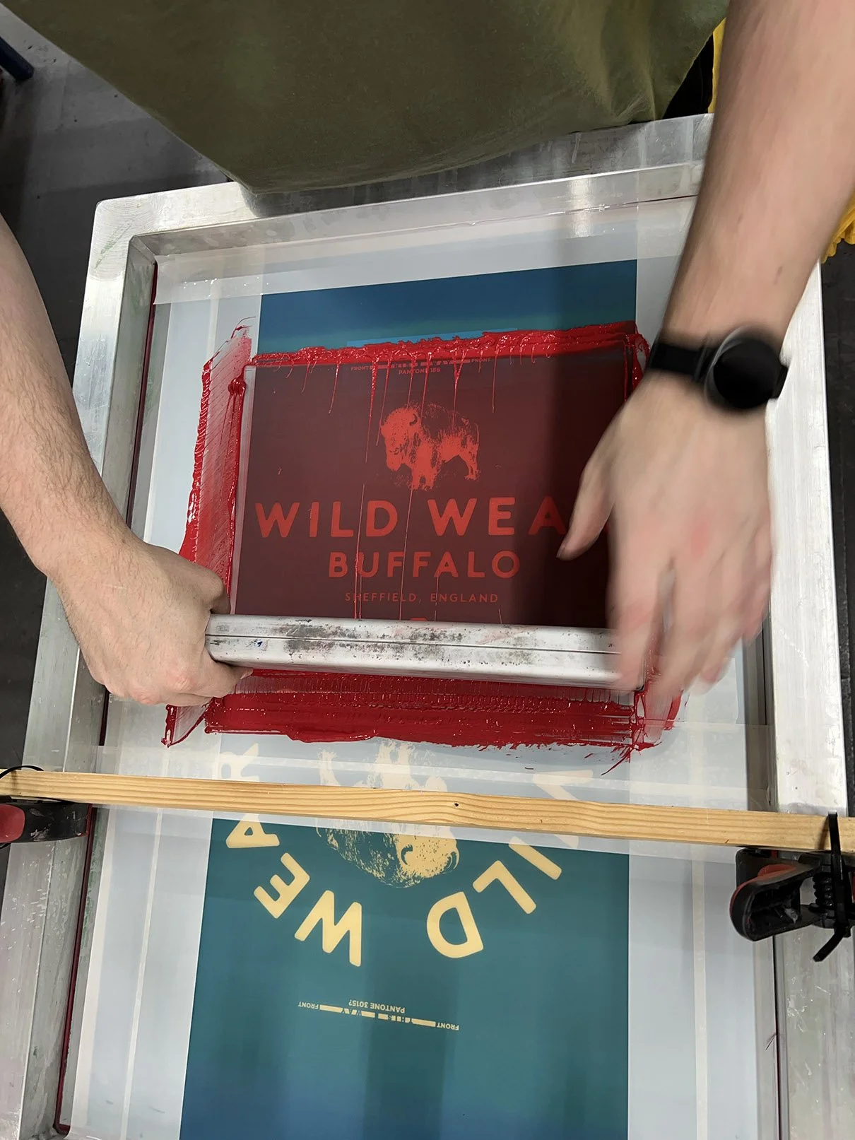 A person is screen printing a sign that reads "WILD WEAR BUFFALO SHEFFIELD, ENGLAND" onto a red screen, using a printing frame and a squeegee. The workspace includes a large print on a teal background with a logo.