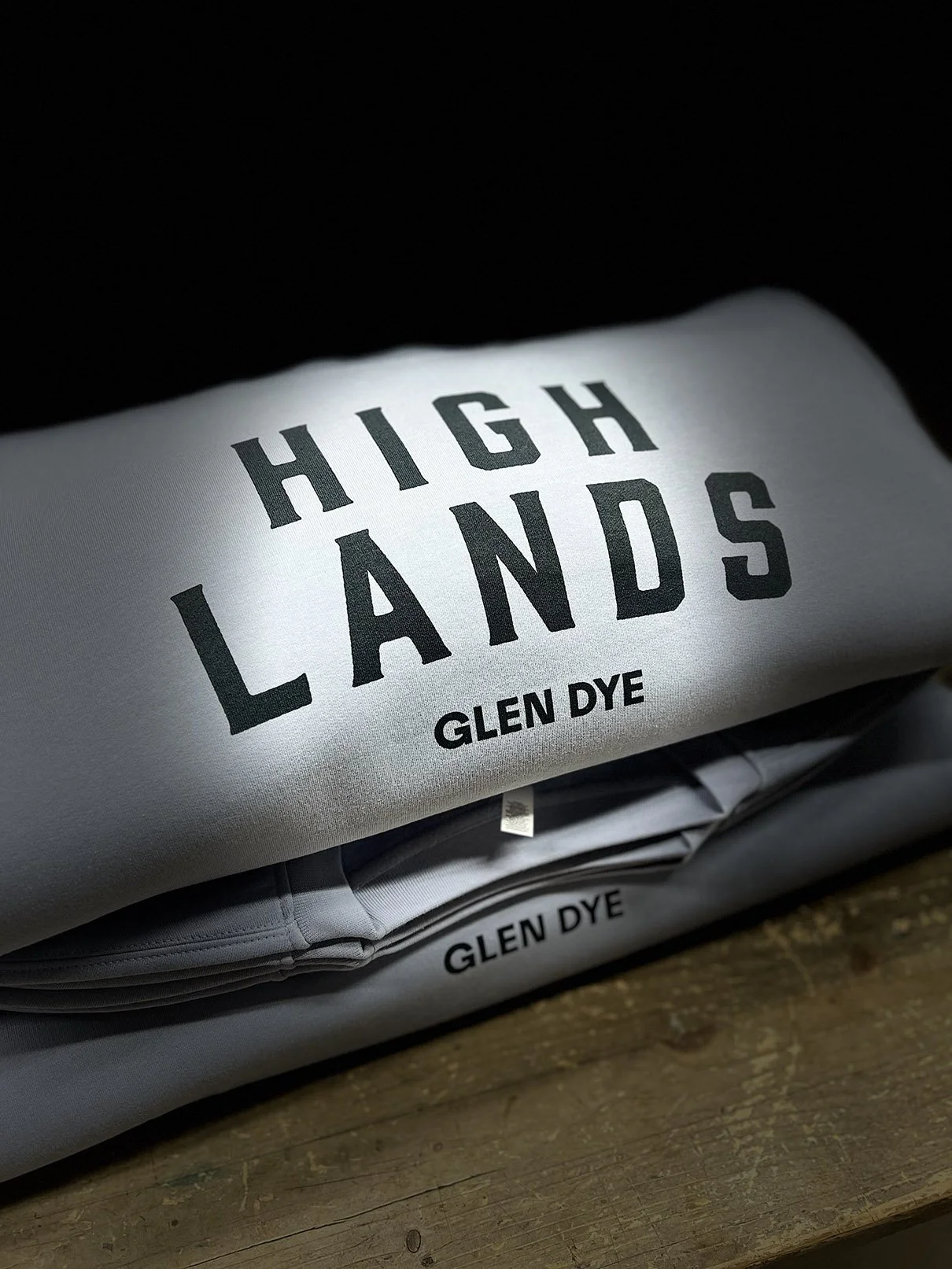 A folded heather grey sweatshirt of high-end clothing from Glen Dye with the brand name printed in black, placed on a wooden surface.