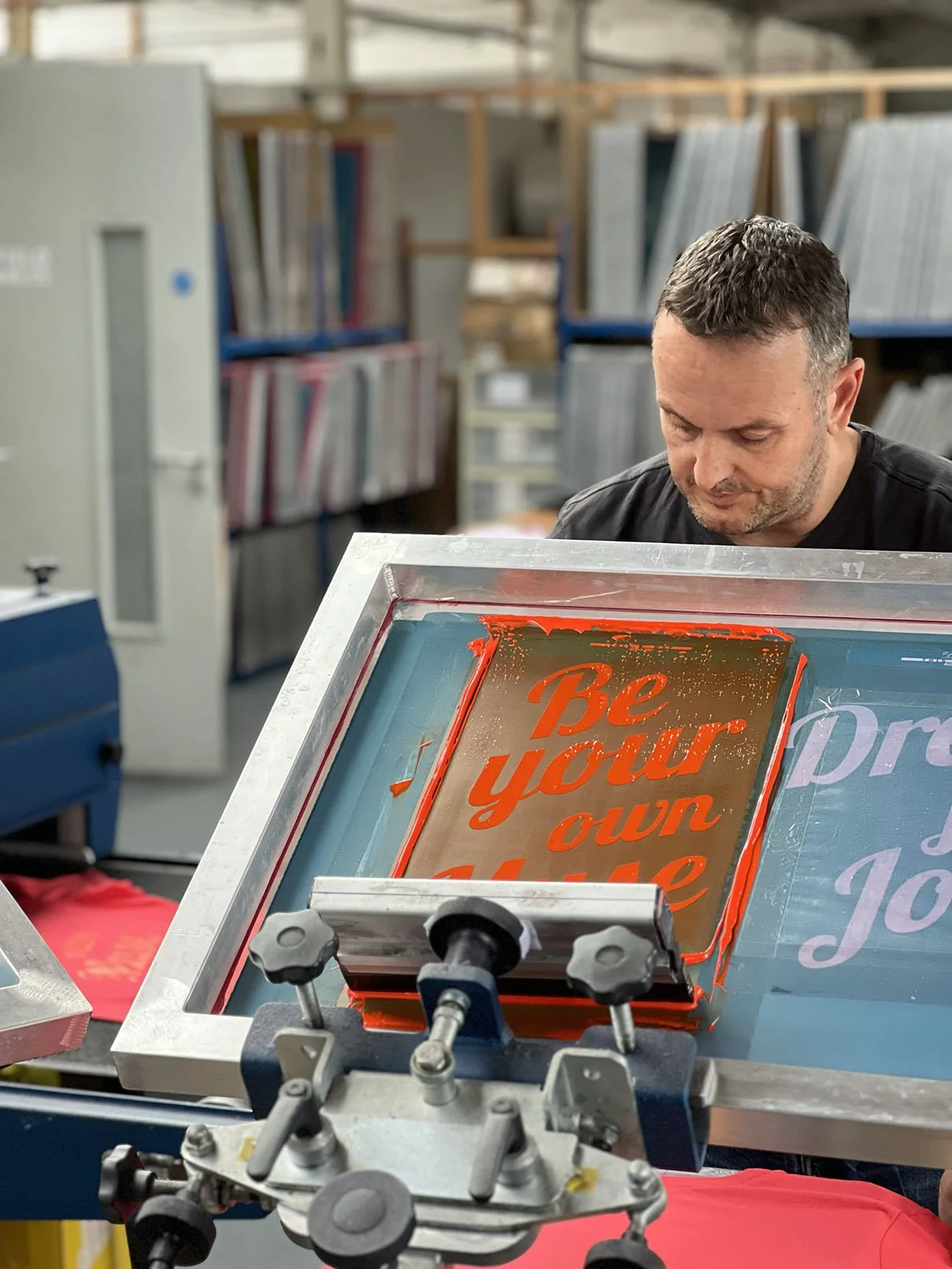 A man working on a screen printing press with a framed design that says 'Be your own'. The workspace contains shelves of books and materials.