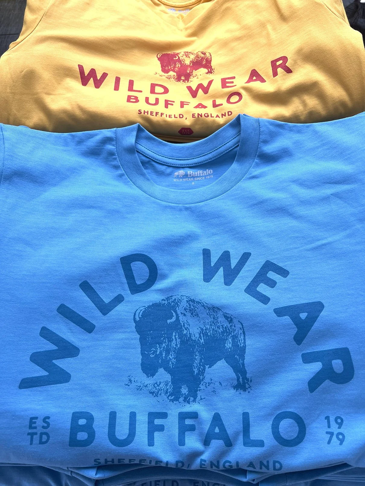 Two T-shirts, one yellow with red and pink text that reads "Wild Wear Buffalo" and a buffalo graphic, the other blue with a buffalo graphic and text that reads "Wild Wear Buffalo" and "Sheffield, England." for Buffalo Systems