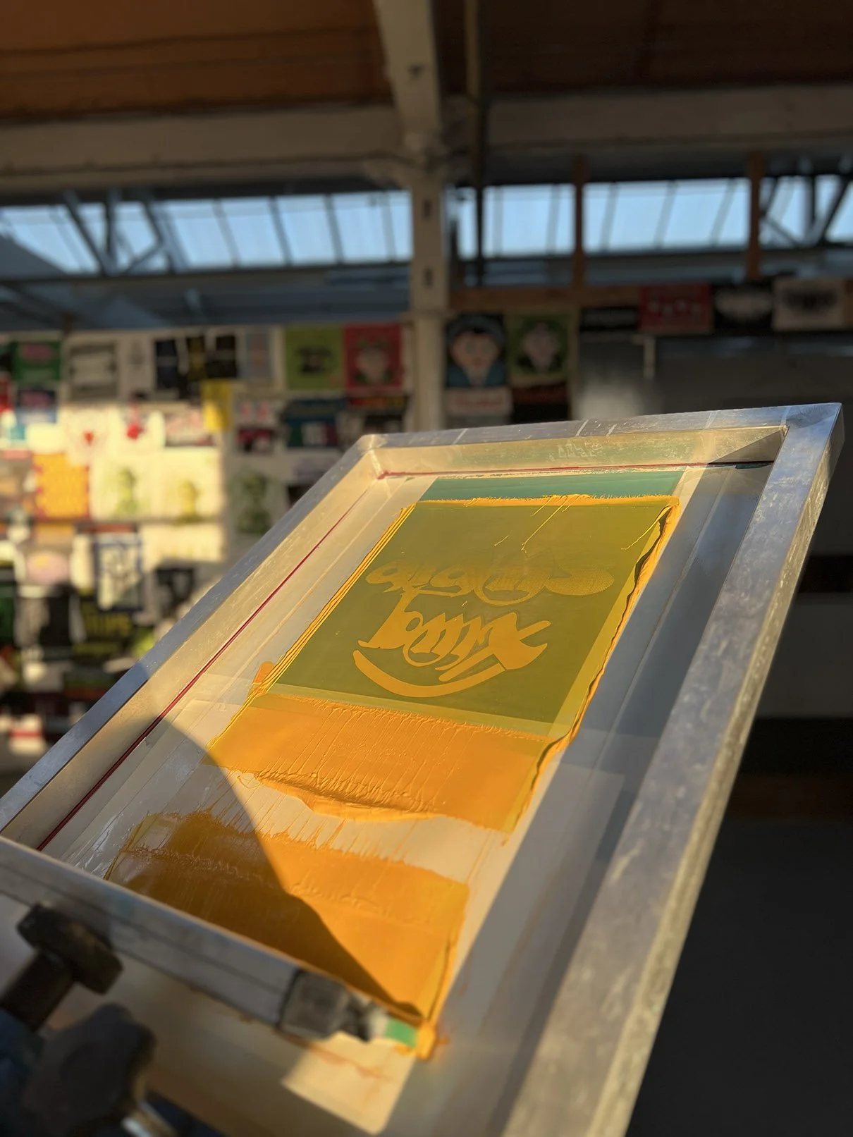 Printing screen with yellow ink on a silk screen printing frame, inside a warehouse or workshop with various signs and products in the background.