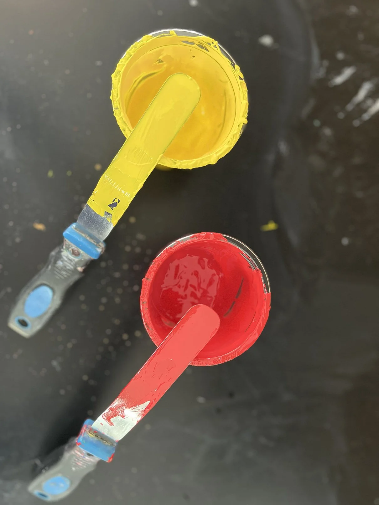 Top-down view of two paint containers, one yellow and one red, with paintbrushes dipped inside and resting on a black surface.