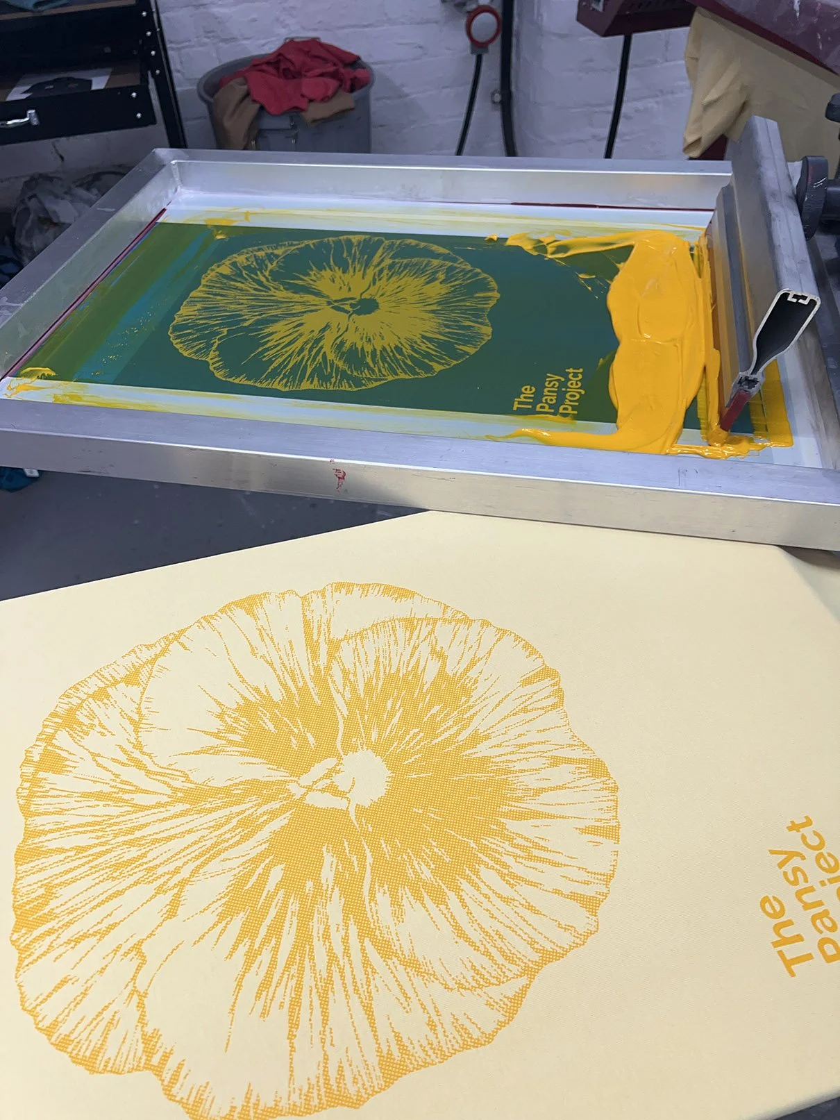 A screen printing setup for a yellow line illustration of a pansy for  'The Pansy Project' showing a silk screen, and a print with the same design on a t-shirt.