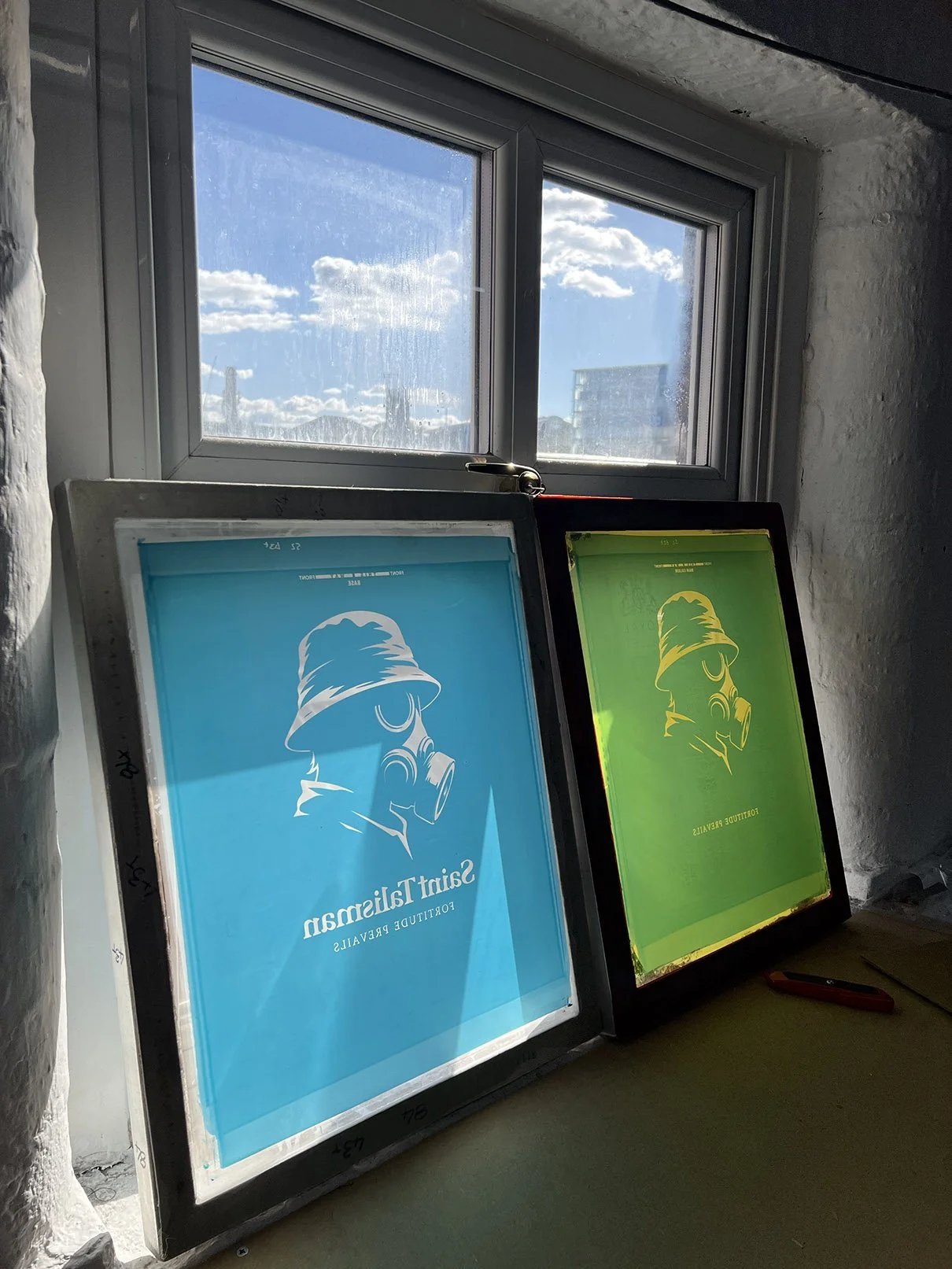 Two illuminated silkscreens leaning against a window, one blue with an illustration of a person wearing a gas mask and a bucket hat, captioned 'Saint Talisman' and the other green with the same illustration, both displaying related graphics.