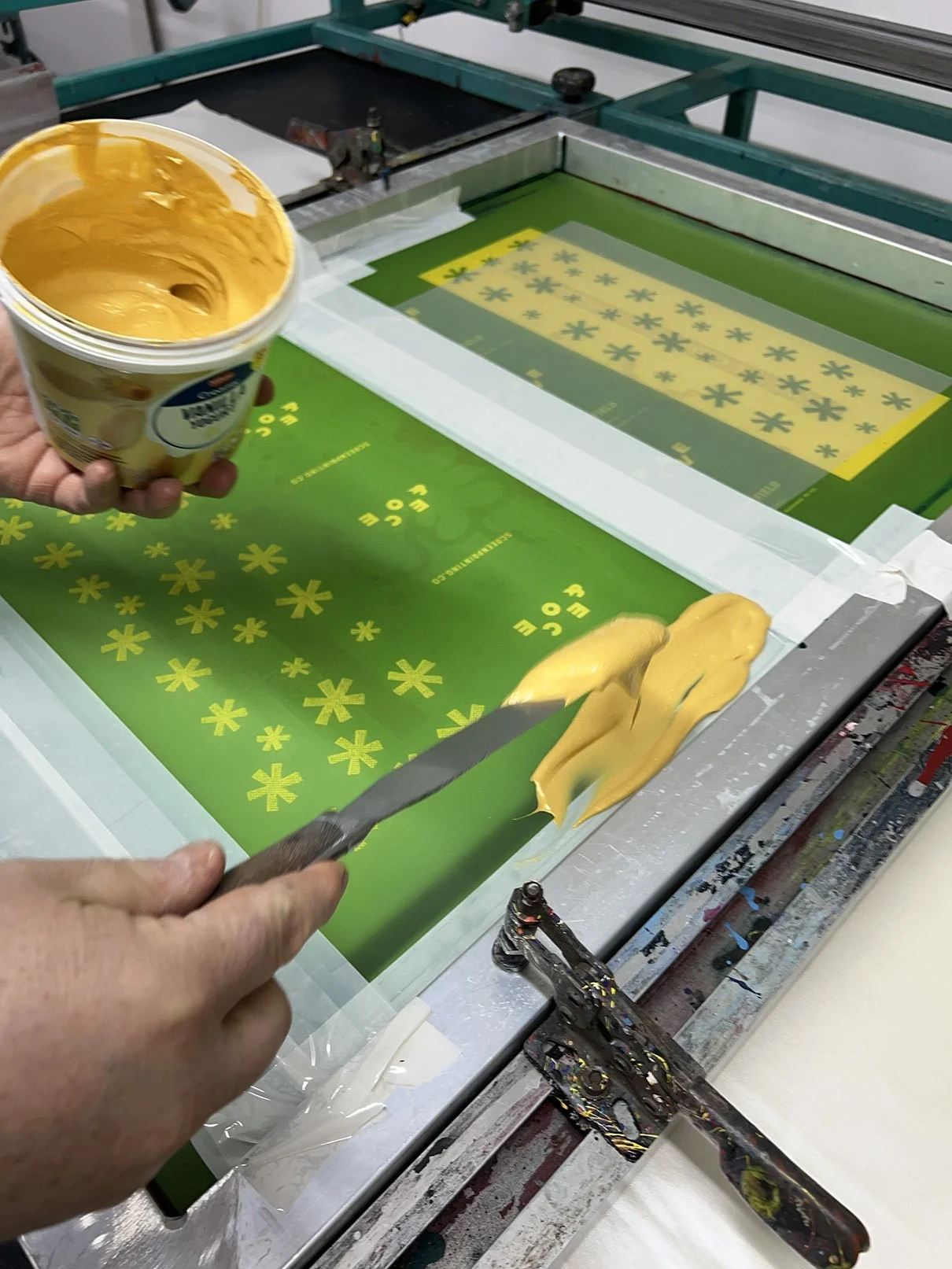 A person prints a yellow flower pattern onto a green sheet using a screen printing frame. They hold a yellow ink tub in one hand and use a squeegee to spread ink over the screen.