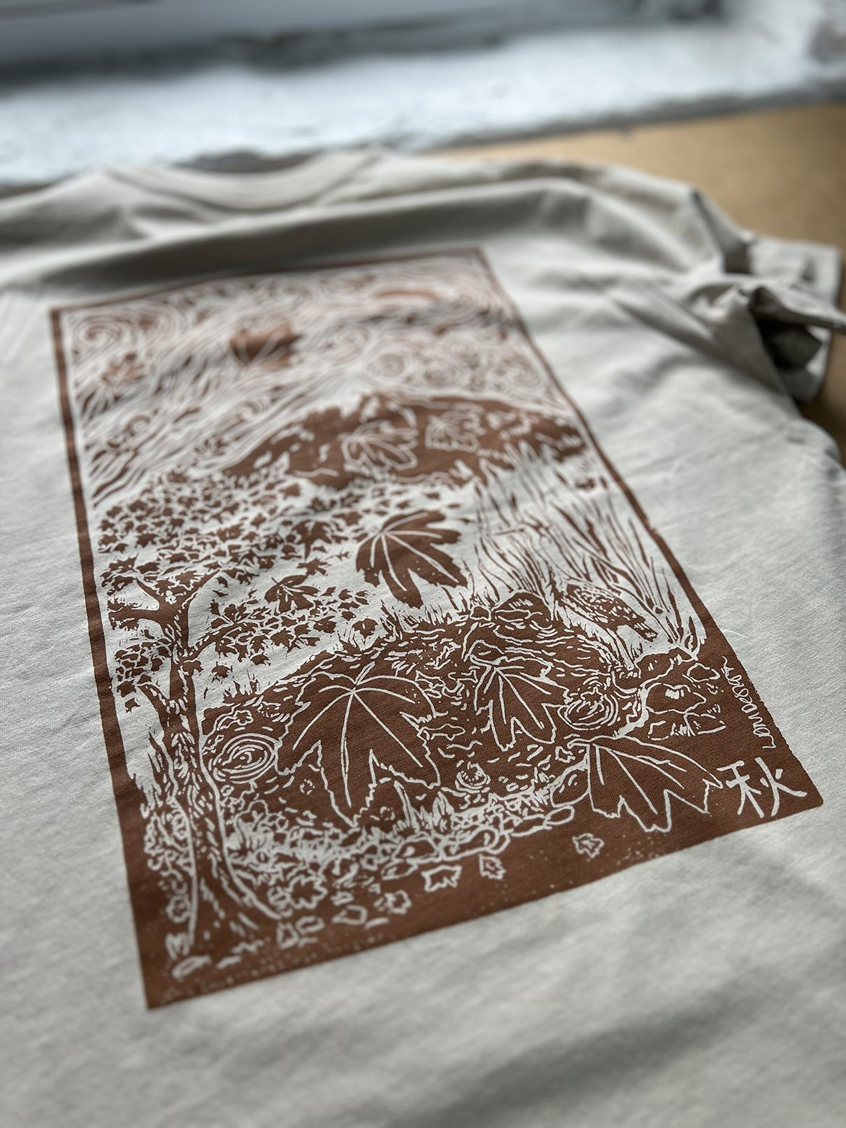 Close-up of a natural coloured organic cotton, ethically made t-shirt with a brown illustration of nature, including leaves, trees, and a bear, printed on it for LDM Design.