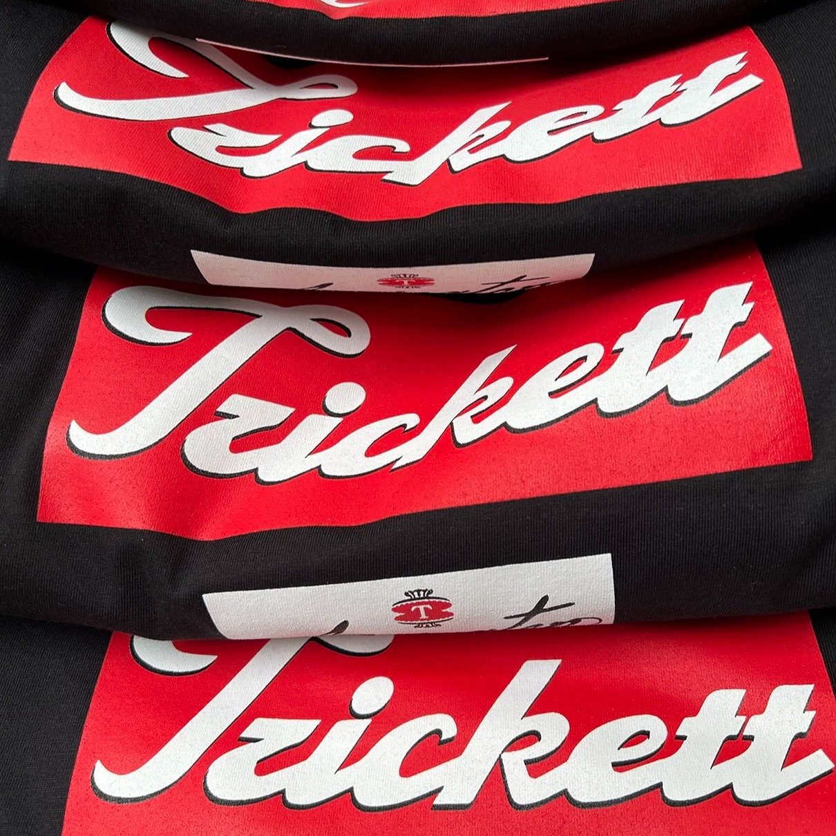 Stacked black t-shirts with screen printed red and white 'Trickett' logo.