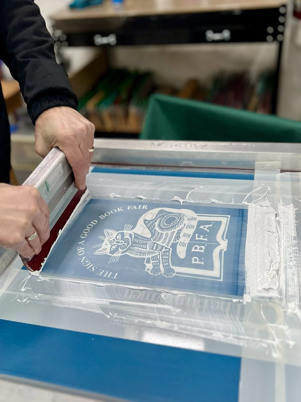 A person using a squeegee to screen print a book cover for the PBFA book fair, featuring a cat logo and the text 'The Sign of a Good Book Fair'.
