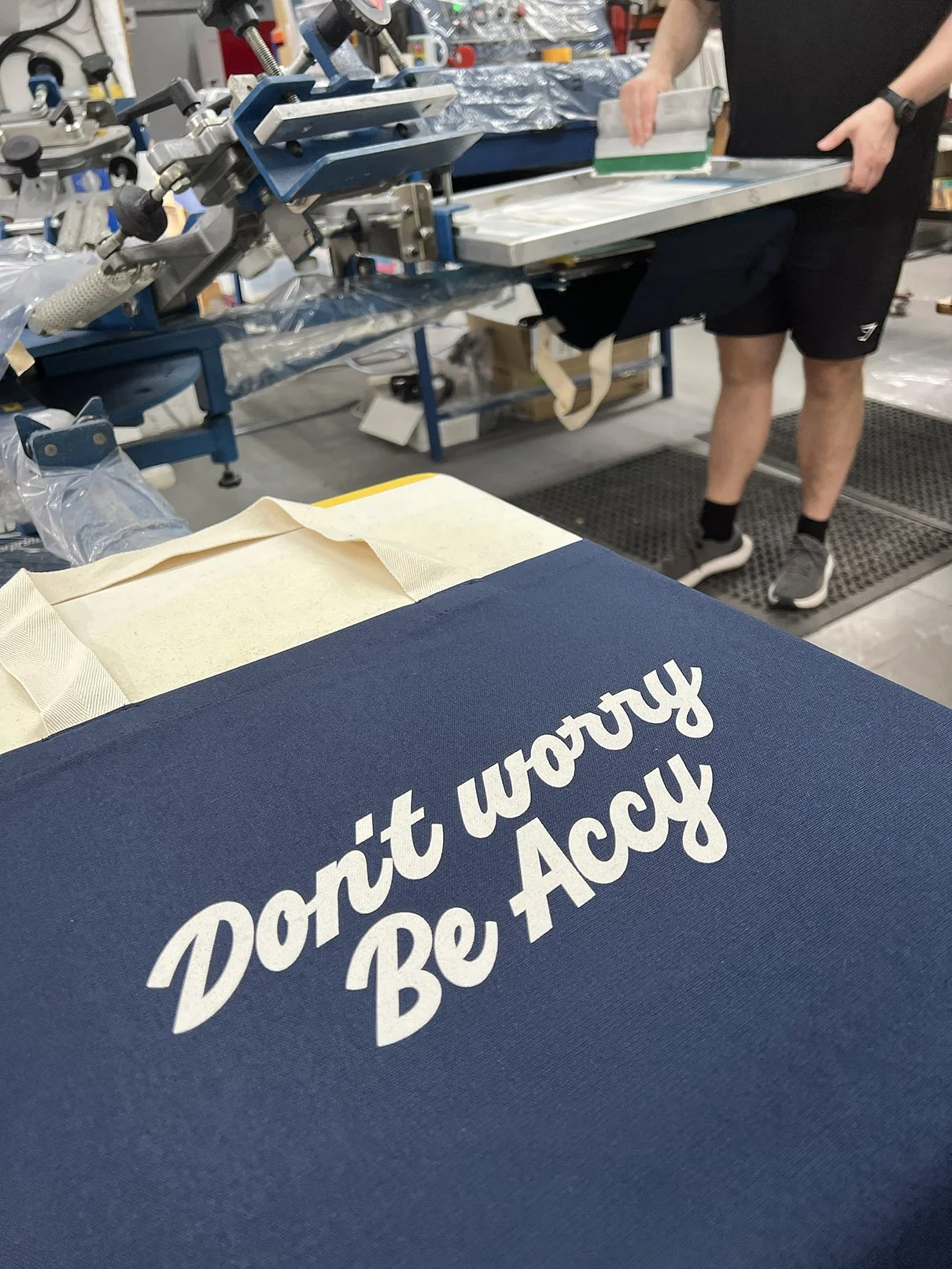 A blue tote bag with the phrase 'Don't worry Be Accy' screenprinted in white ink, placed on a screenprinting machine.