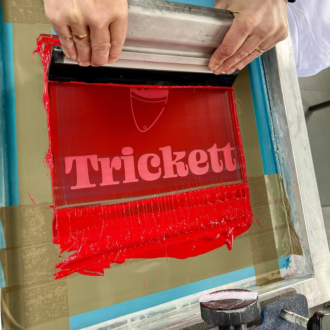 A person is pressing a red screen with the word 'Tricket' on it, used in screen printing, with red ink surrounding the edges.