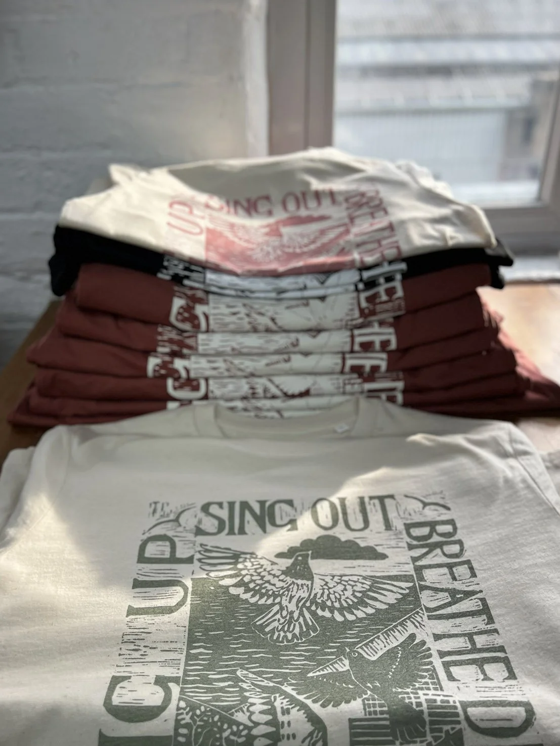 Stacked folded T-shirts with a design featuring a bird and the text "The Sing Out Brigade," placed on a wooden surface near a window.