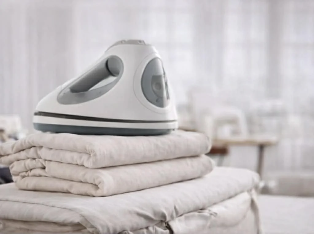 An iron resting on folded light-colored blankets on an ironing board.