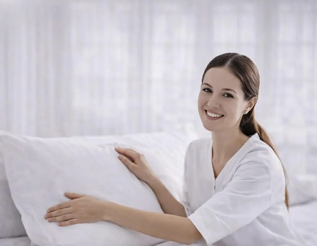 A young woman with brown hair in a ponytail, smiling and wearing a white shirt, tidying a white pillow in a bright bedroom.