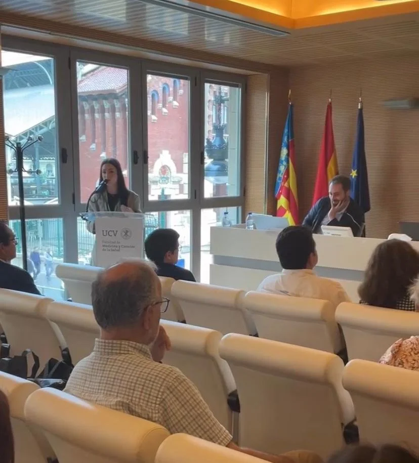 Una conferencia en una sala con varias personas, una mujer hablando en un podio con una micrófono, y un hombre sentado en el escritorio tras un micrófono. Detrás, banderas de Valencia, España, y la Unión Europea.