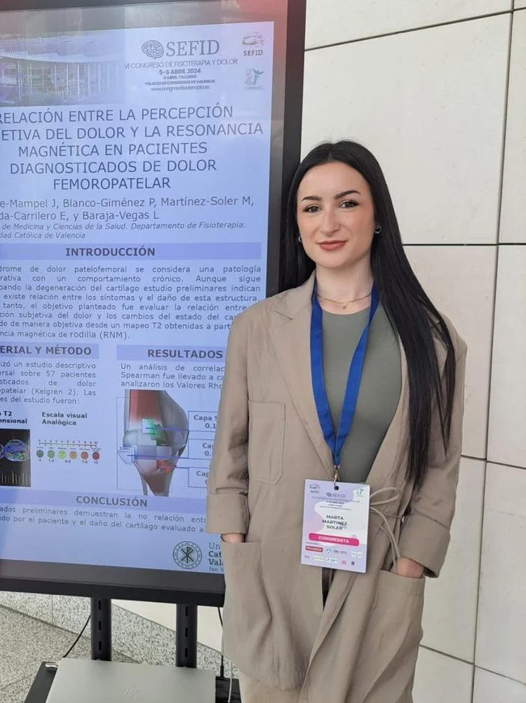 Una mujer con cabello largo y oscuro, vestida con un saco beige, de pie frente a un cartel en una conferencia científica. El cartel trata sobre la percepción del dolor y la resonancia magnética en pacientes con dolor femoropatelar.