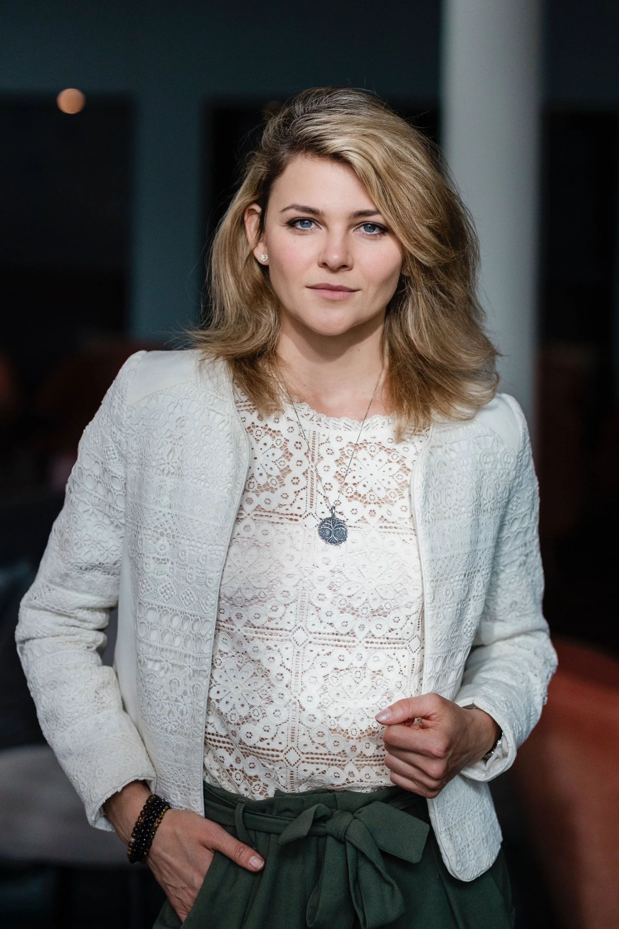 En kvinna med blont, vänner hår och blå ögon står mot en mörk bakgrund, klädd i en vit blus och matchande blazer, bär armband och ett halsband.