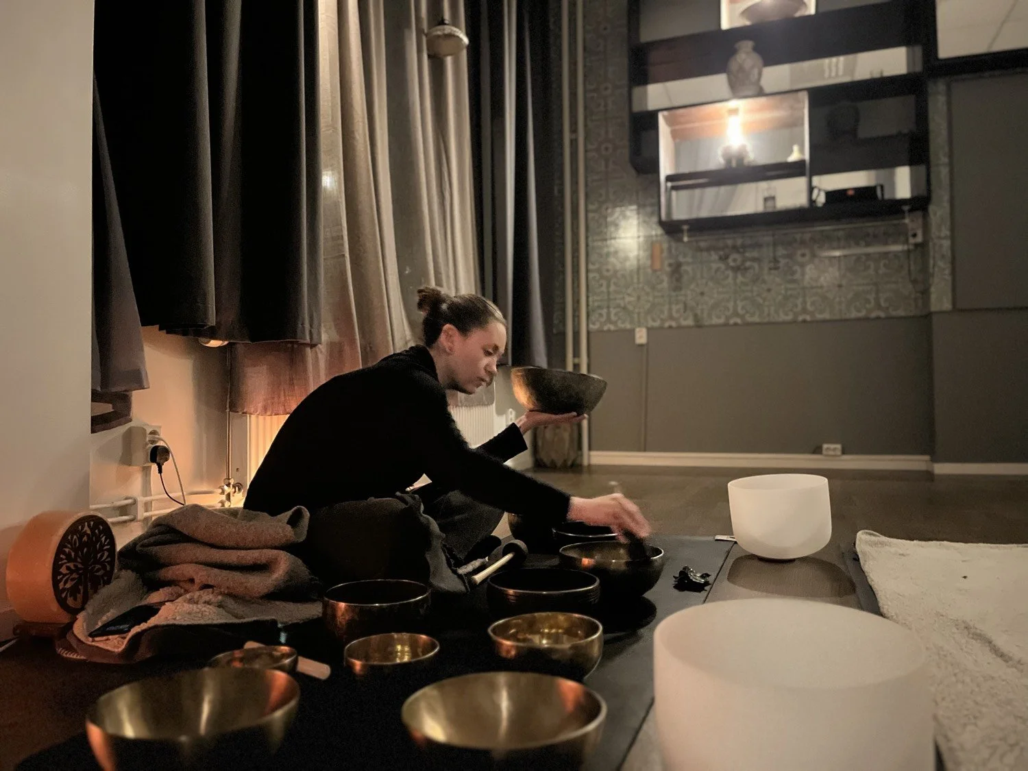 En person som spelar på kristallskålar i ett rum med mörka väggar och gardiner, på en låg bord med olika musikinstrument och energikristaller.