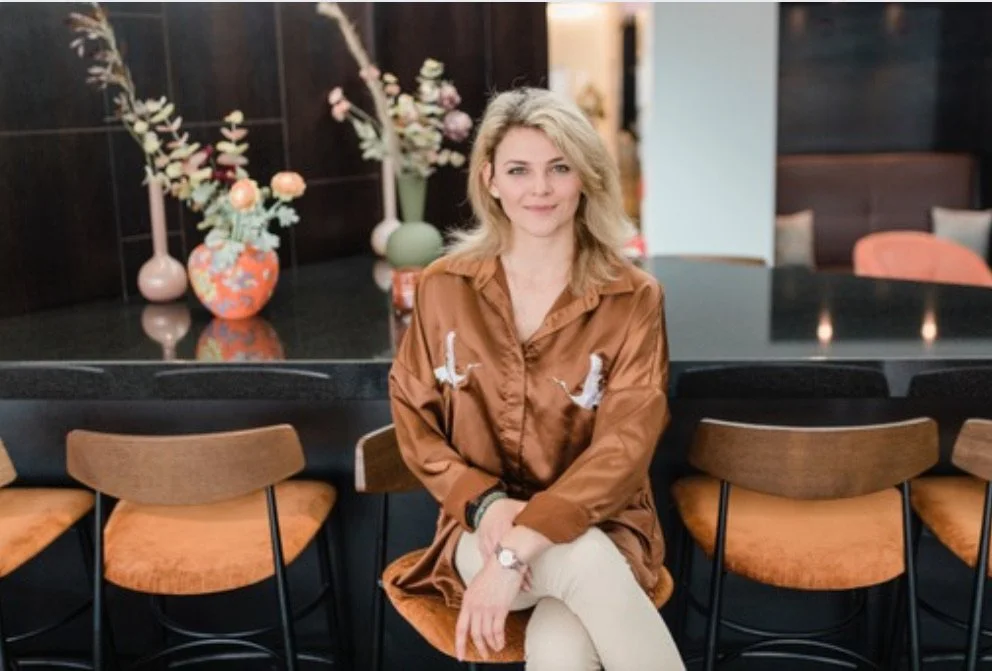 En kvinna med blont, lockigt hår sitter på en barstol vid ett svart köksbord i ett modernt interiör med blommor i bakgrunden.