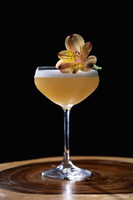 A cocktail glass with a yellowish drink, garnished with a yellow and pink flower, on a wooden surface against a black background.