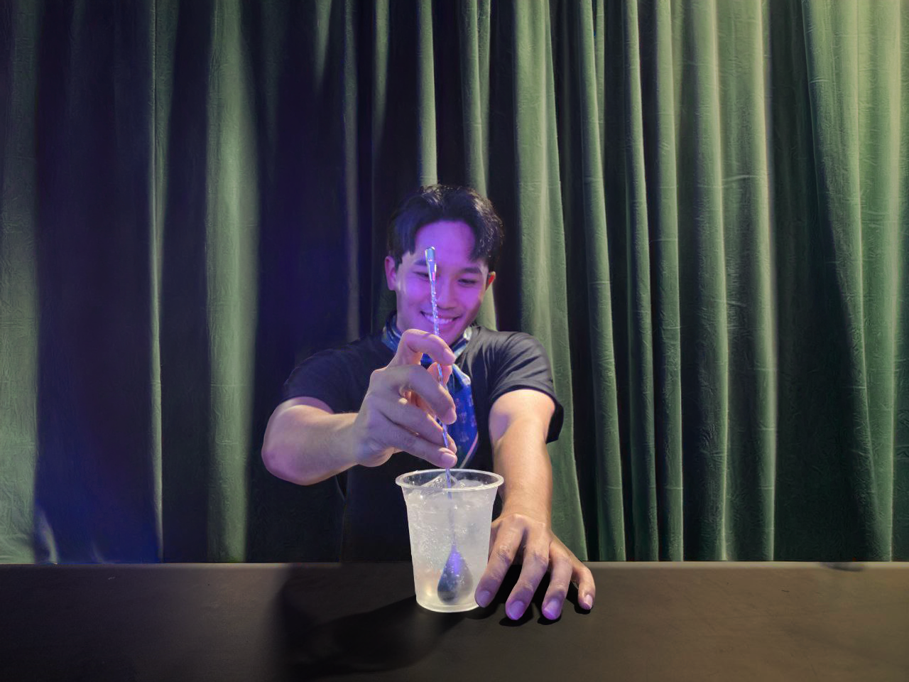A young man smiling as he stirs a transparent cup with a spoon, with a popsicle stick in his hand. The background features dark green curtains.
