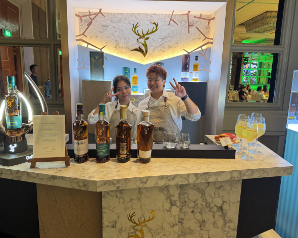 Two smiling bartenders behind a marble bar counter with bottles of liquor, a drink menu, and cocktails, in a modern bar with decorative lighting and a deer head logo on the back wall.