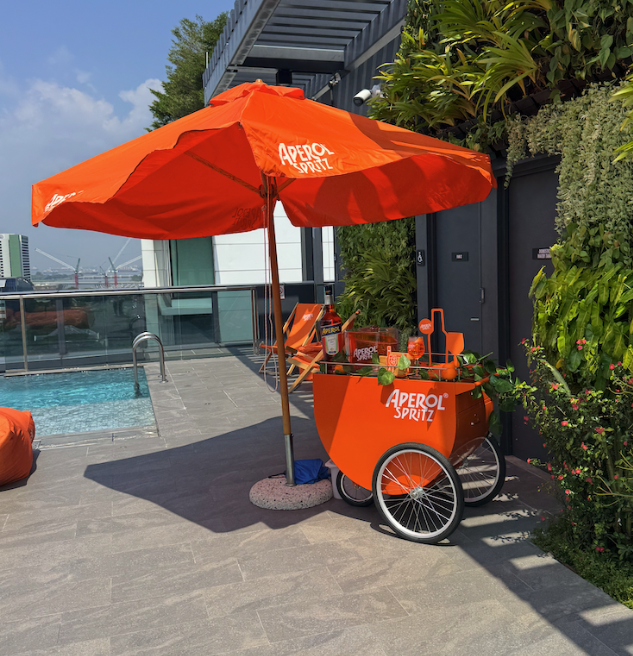 A bright orange Aperol Spritz cart with matching umbrella on a rooftop patio next to a pool, with some greenery and city buildings in the background.