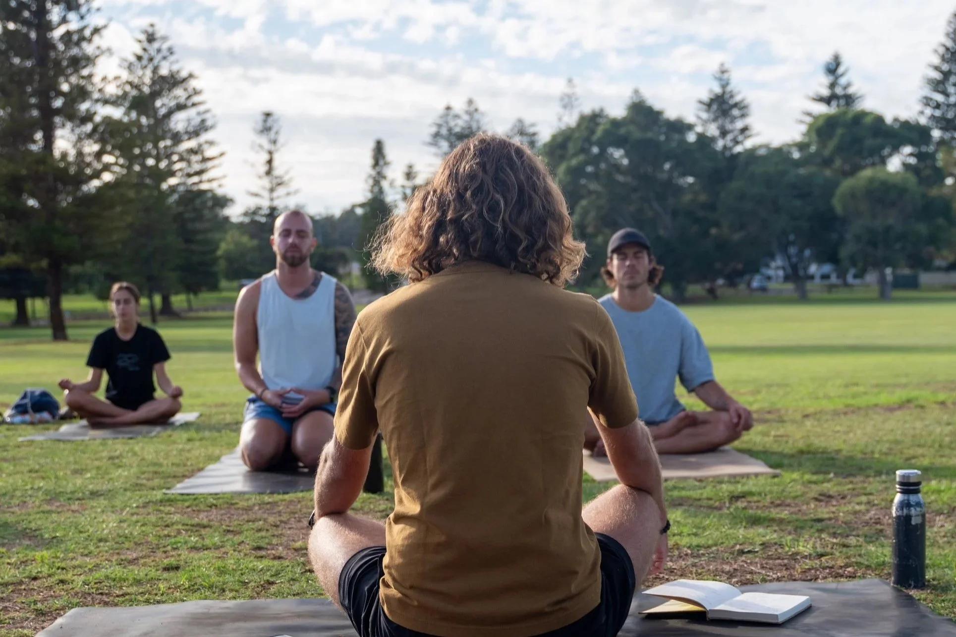 A group of four young adults participating in an outdoor yoga or meditation session in a park, sitting cross-legged on mats with trees and a blue sky in the background.