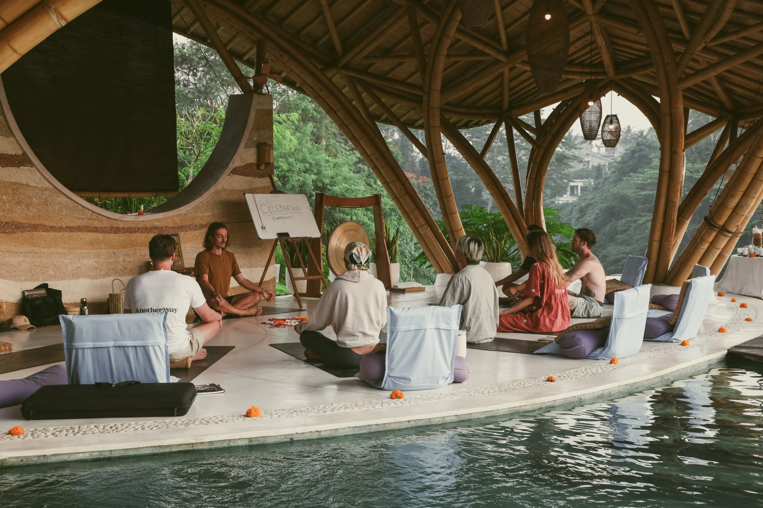 A group of people participating in a meditation or yoga session in an open-air bamboo structure with a view of lush greenery outside.