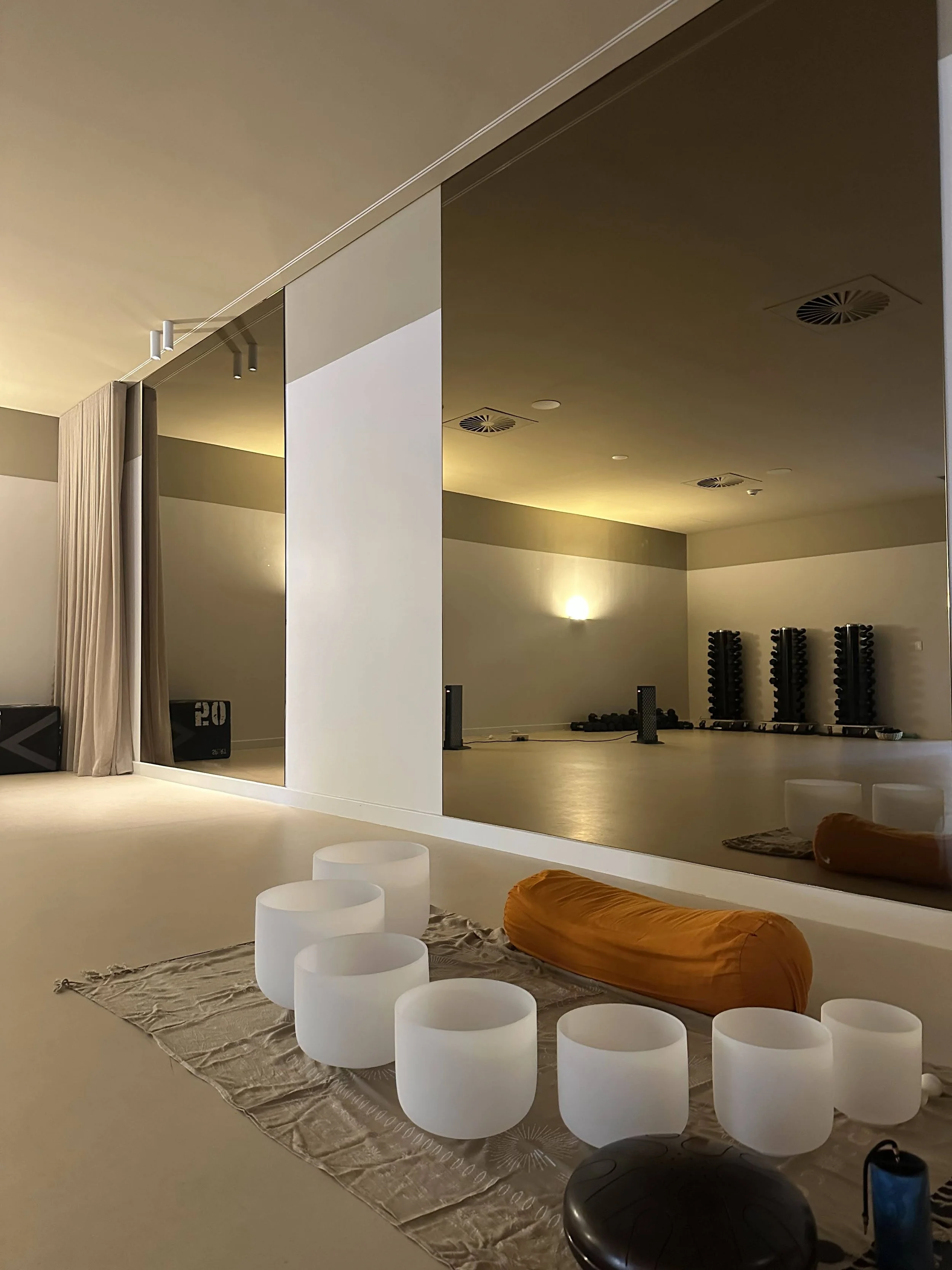 A peaceful yoga or meditation studio with glowing white bowls, an orange bolster, and a mirror reflecting the room with dumbbells and weights in the background.