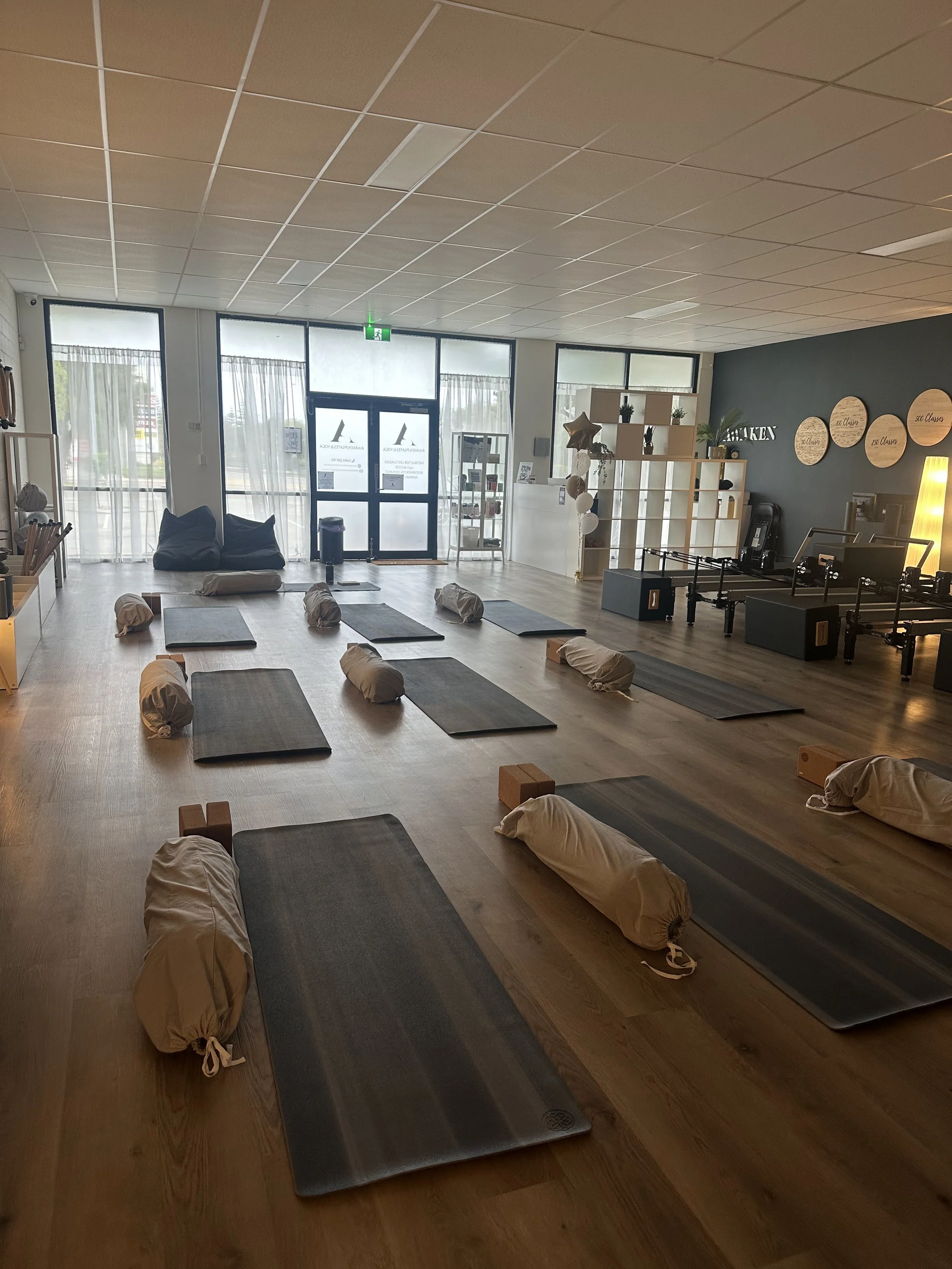 Empty yoga or pilates studio with black mats, wrapped bolsters, wooden blocks, and soft bean bags arranged on a wooden floor, with large windows at the front and a blue accent wall with circular decor.