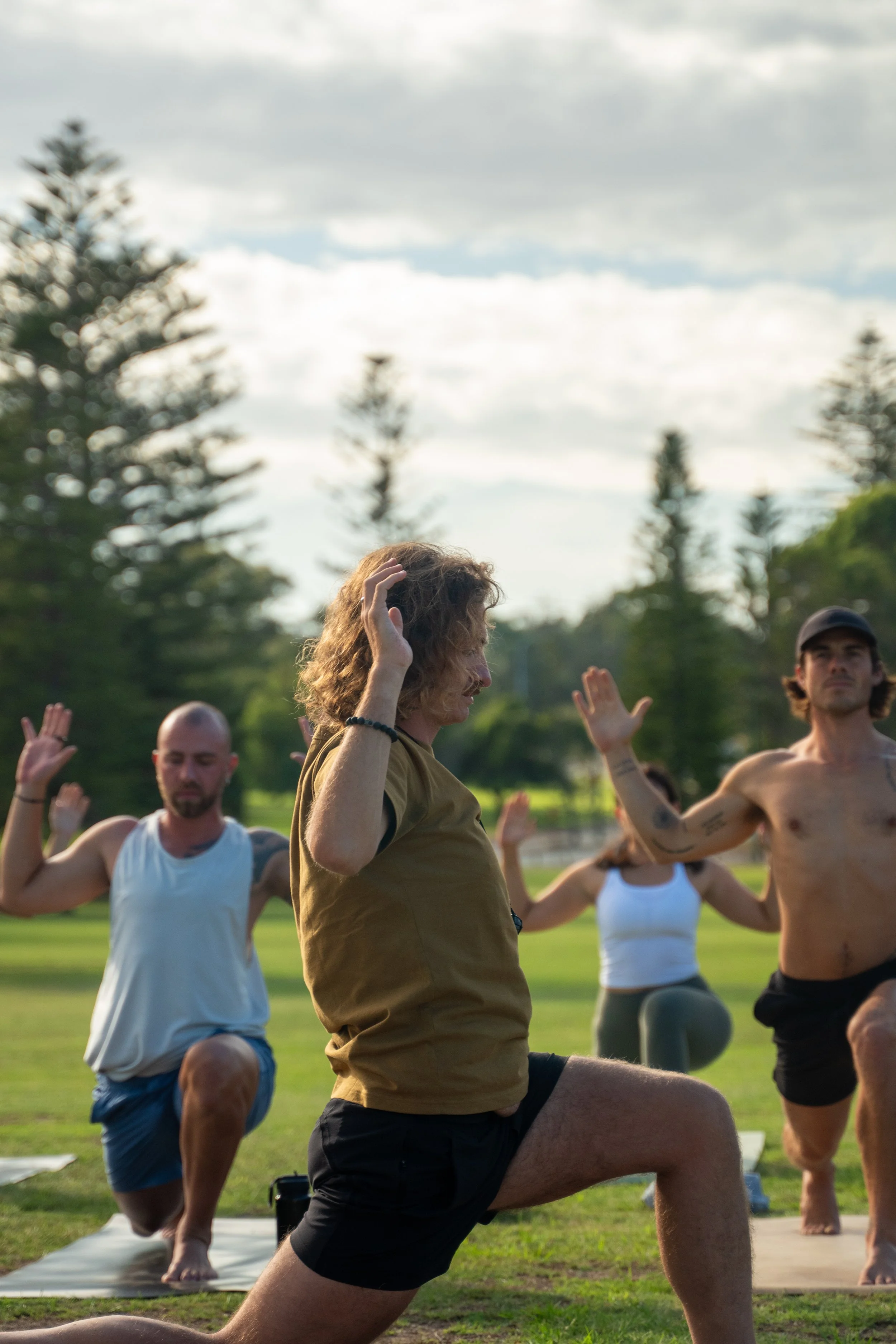 People practicing yoga outdoors on a grassy field with trees and a cloudy sky in the background.