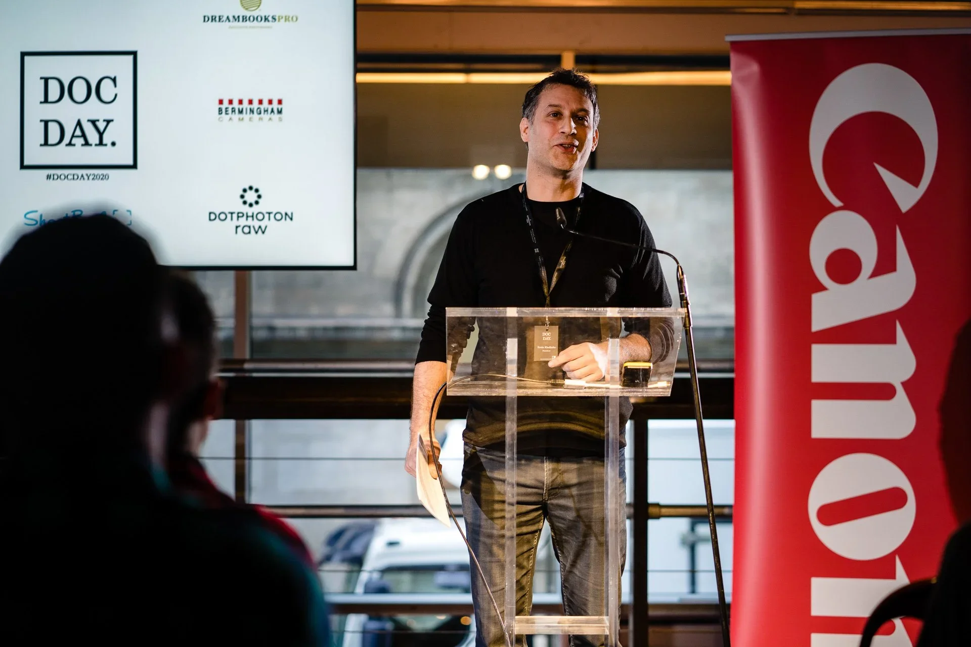 A man giving a presentation at an event called Doc Day, standing behind a clear podium with a microphone, with a large screen displaying logos of sponsors and event hashtags. A red banner with white text is partially visible on the right.