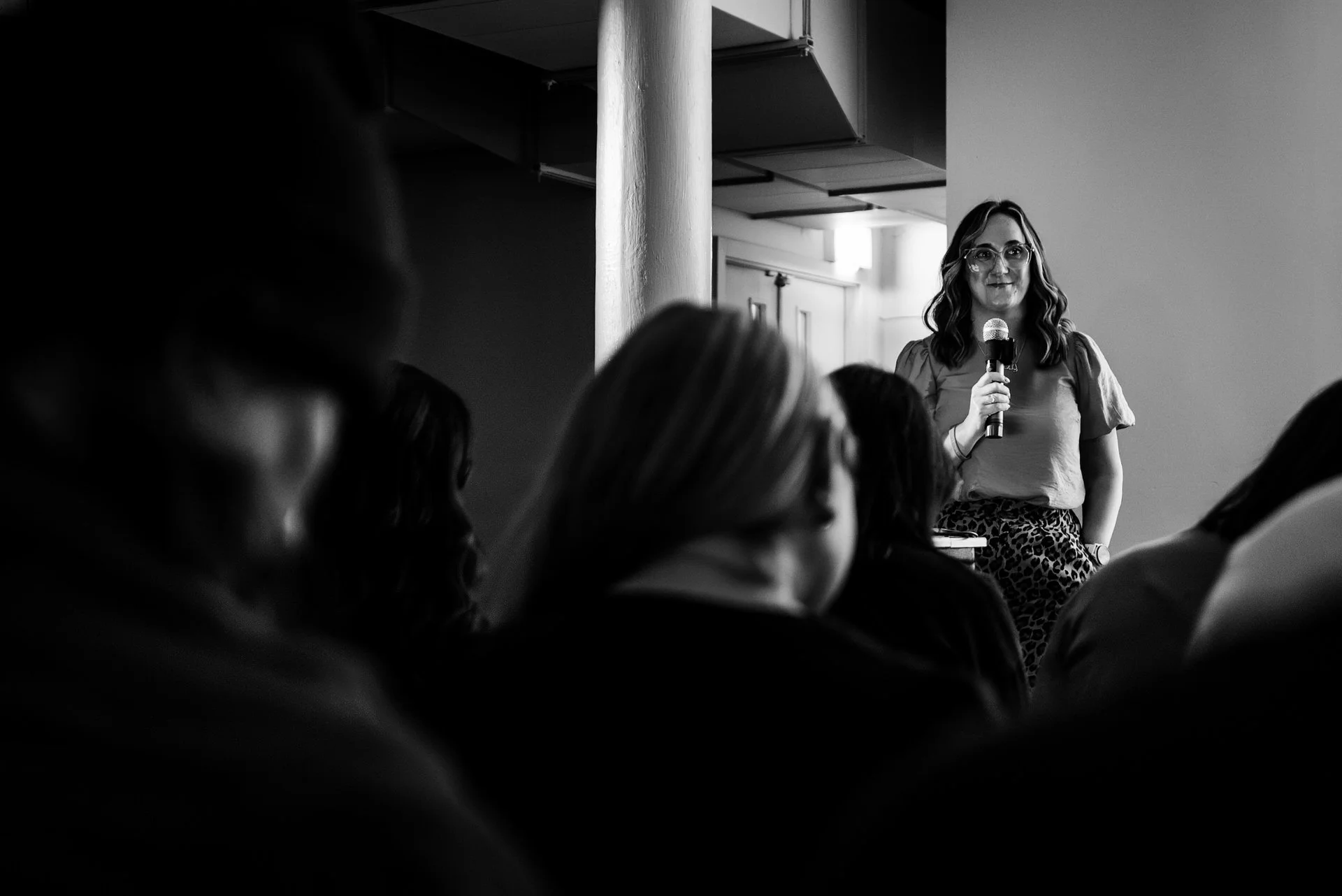 A woman standing and speaking into a microphone in front of an audience in a well-lit room.