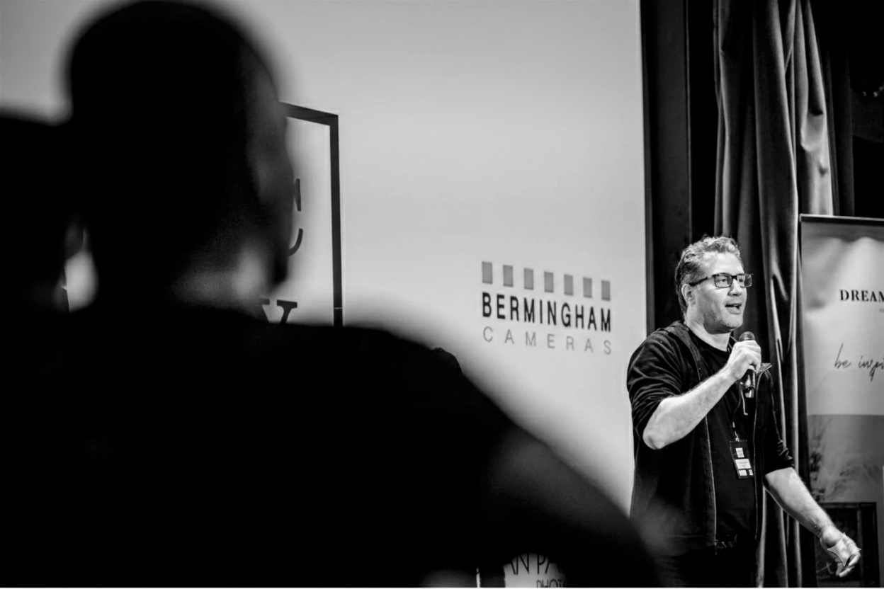 A man with glasses and a beard speaking into a microphone at a presentation, with a large banner and a sign reading 'Bermingham Cameras' in the background, in a black-and-white photo.