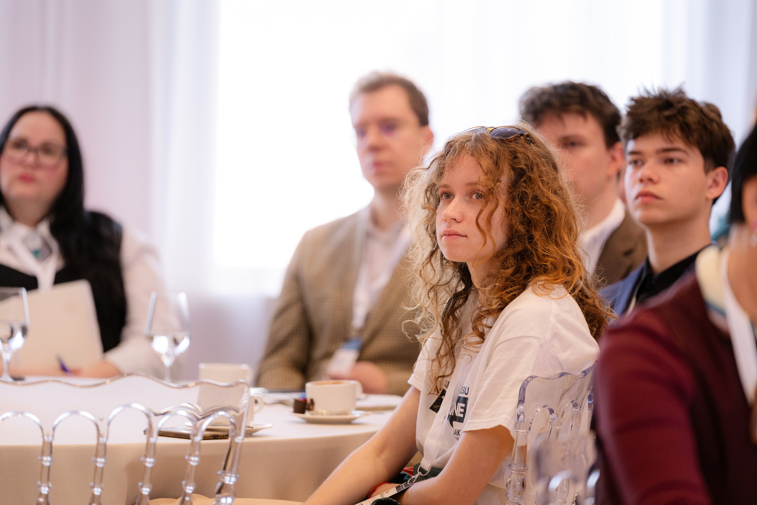 A group of young adults sitting at a conference table, attentively listening during a meeting or seminar, with some having notebooks and coffee cups in front of them.