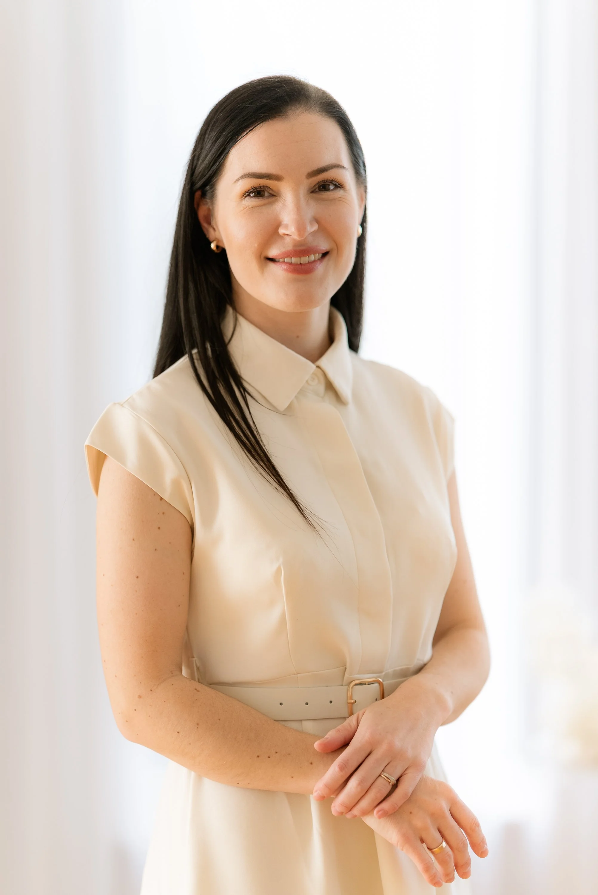 A woman with dark hair wearing a cream-colored dress with short sleeves, standing indoors against a bright background, smiling at the camera.