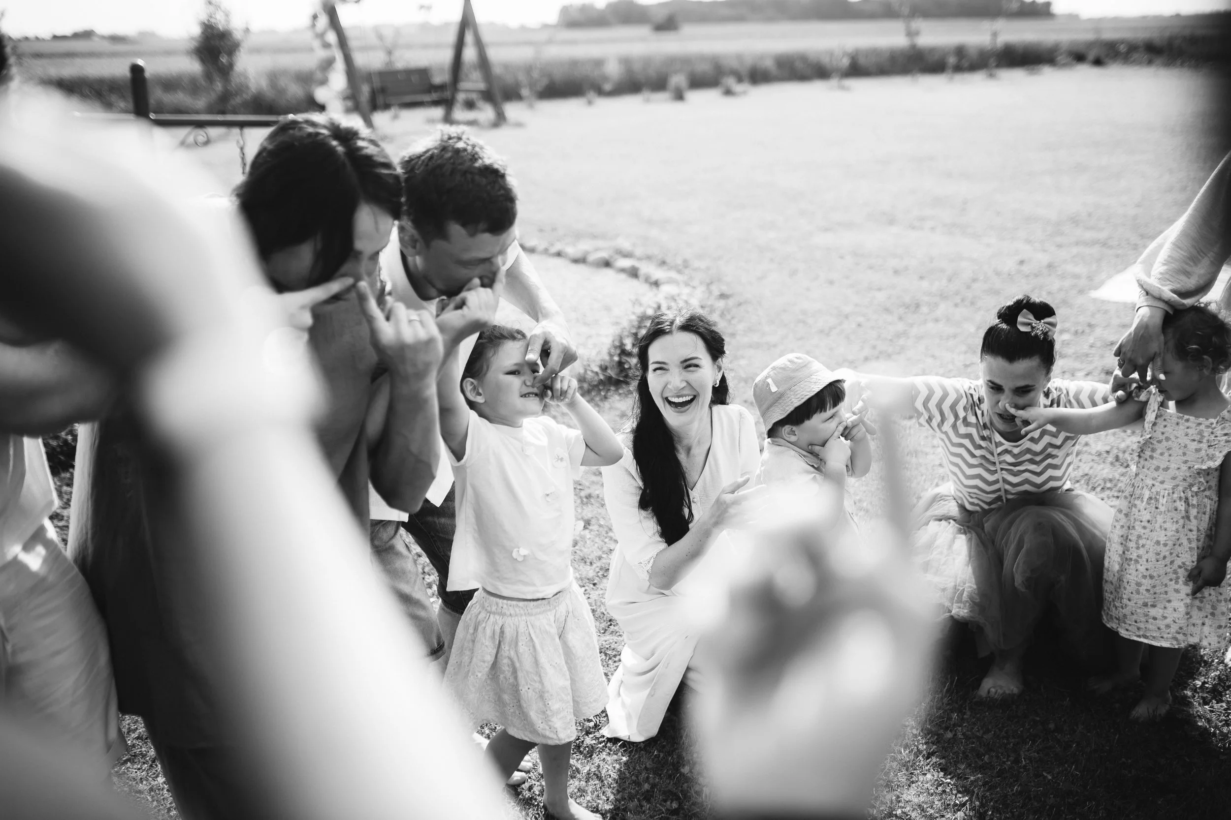 A group of children and women playing a game or engaging in a fun activity outdoors on a grassy field, with some children touching their noses and smiling.