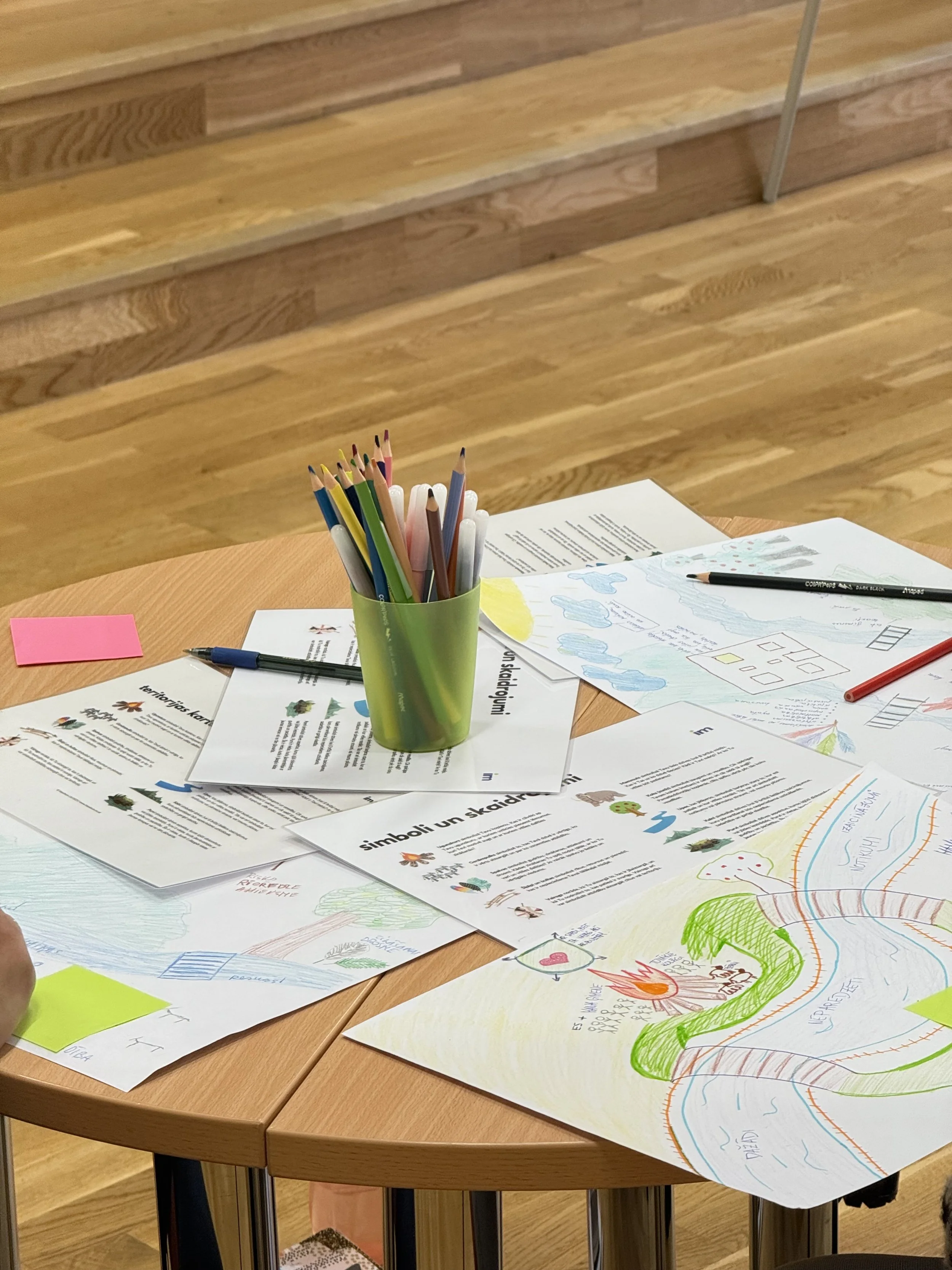 Educational materials and drawings on a wooden table, with a green cup holding colored pencils, colored pens, and various papers, some printed and some hand-drawn.