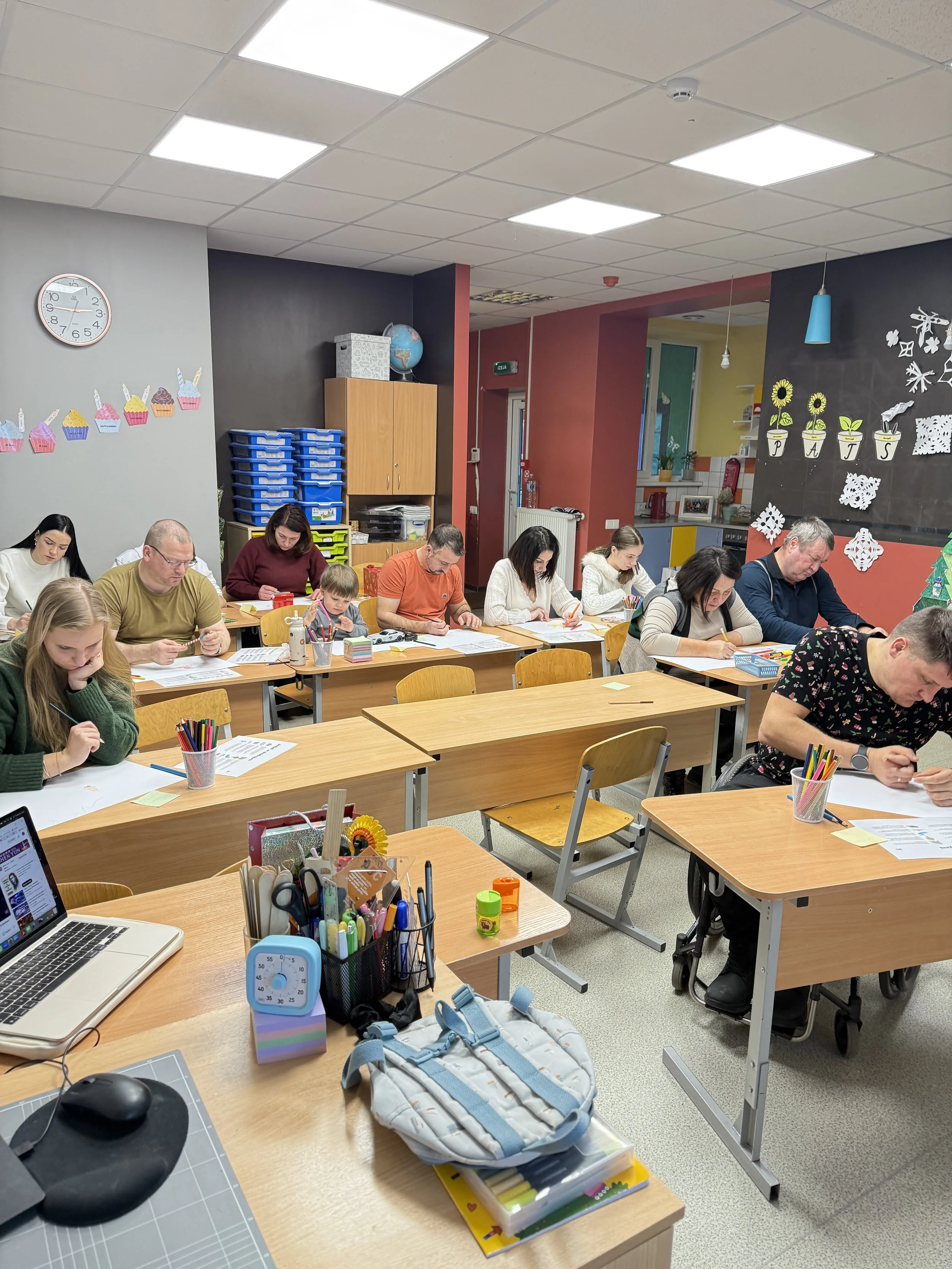 A classroom with adults seated at desks, working on papers. Decorations include cupcakes and potted plants on the walls, and supplies like colored pencils and a laptop on the front desk.