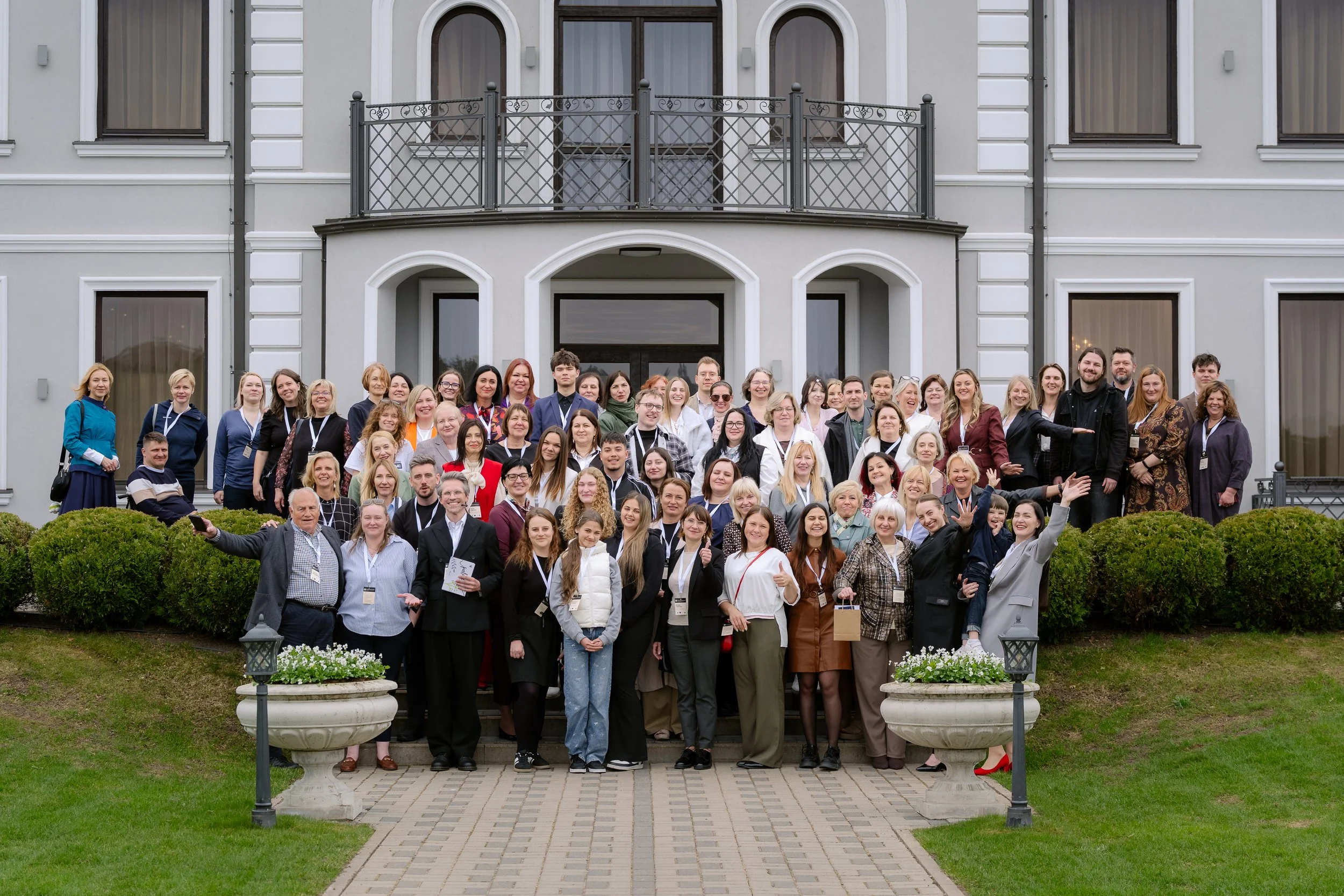 Large group of people posing for a photo in front of a multi-story building with white walls and large windows, some standing on steps and others on the lawn, with greenery and stone planters in the foreground.