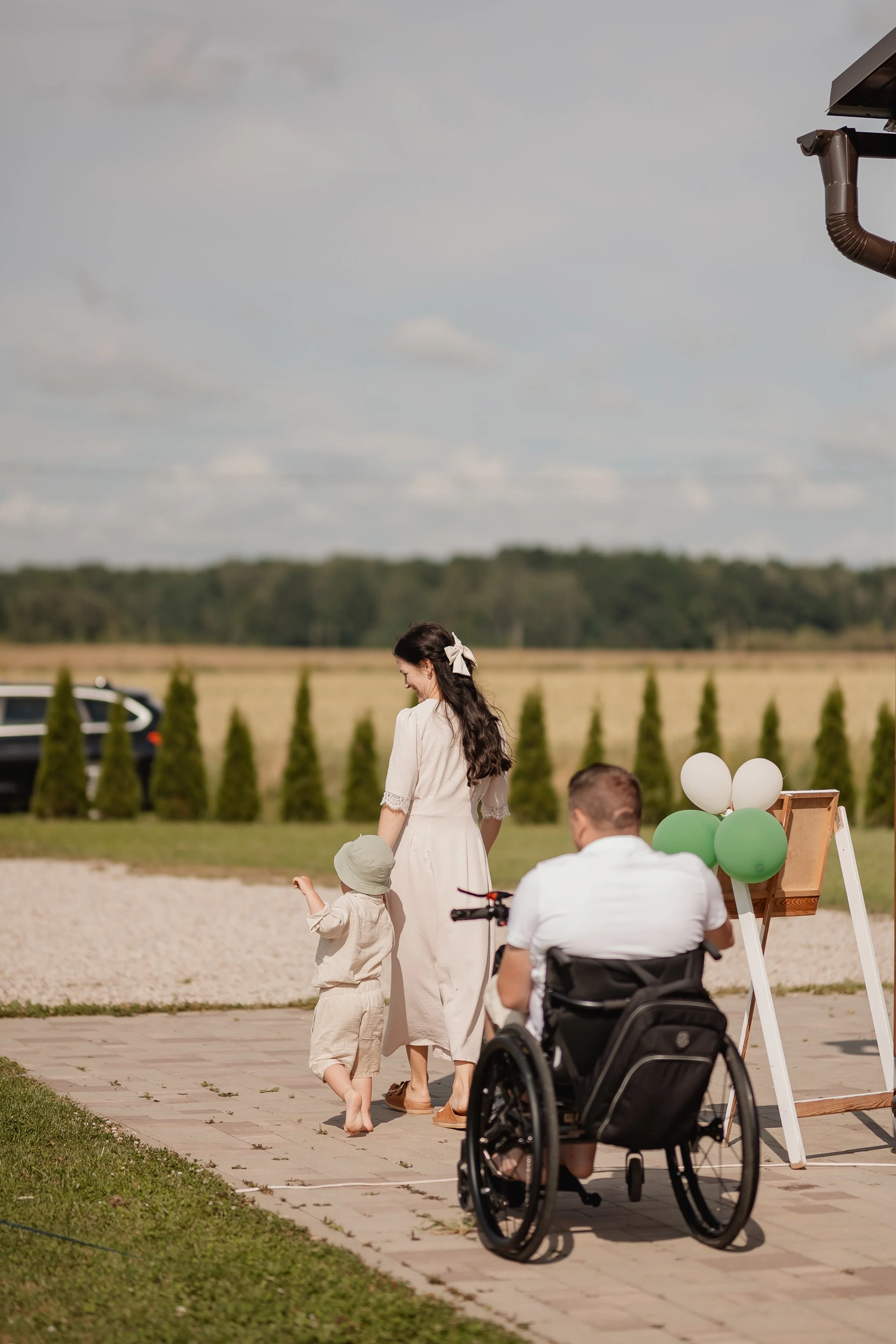 A woman in a white dress holding a child’s hand, with a man in a wheelchair nearby, outdoors during daytime at a celebration with balloons and an easel, against a backdrop of green trees and a cloudy sky.