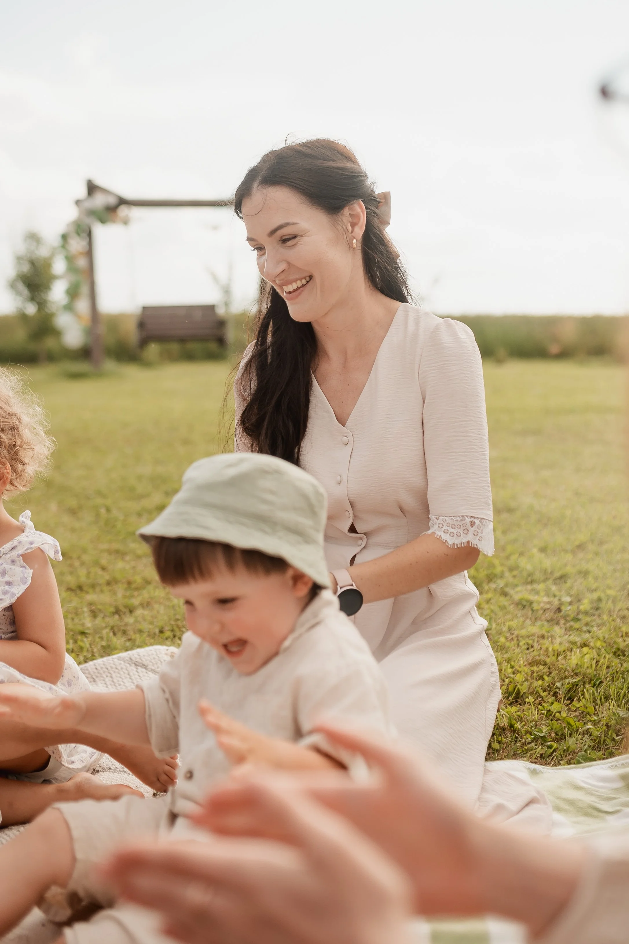 A woman smiling and playing with children outdoors on a grassy field, under a partly cloudy sky.